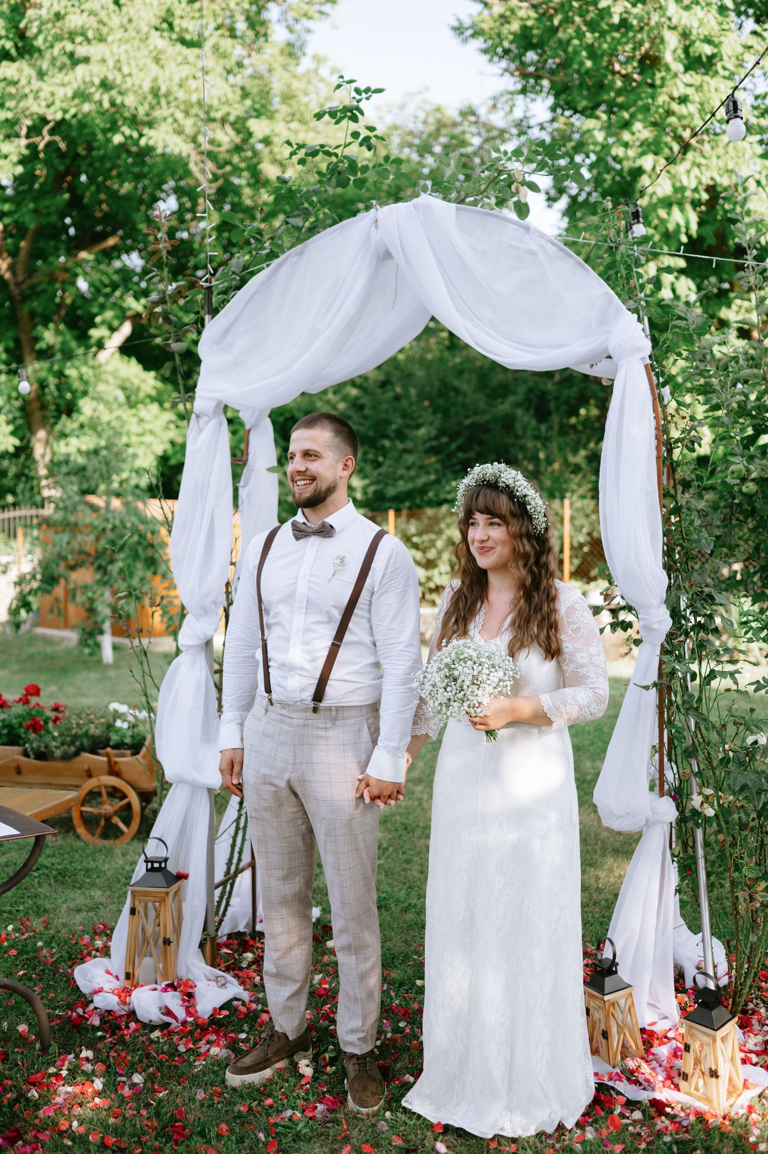 Whimsical outdoor wedding scene with white arch, vintage furniture and scattered rose petals.