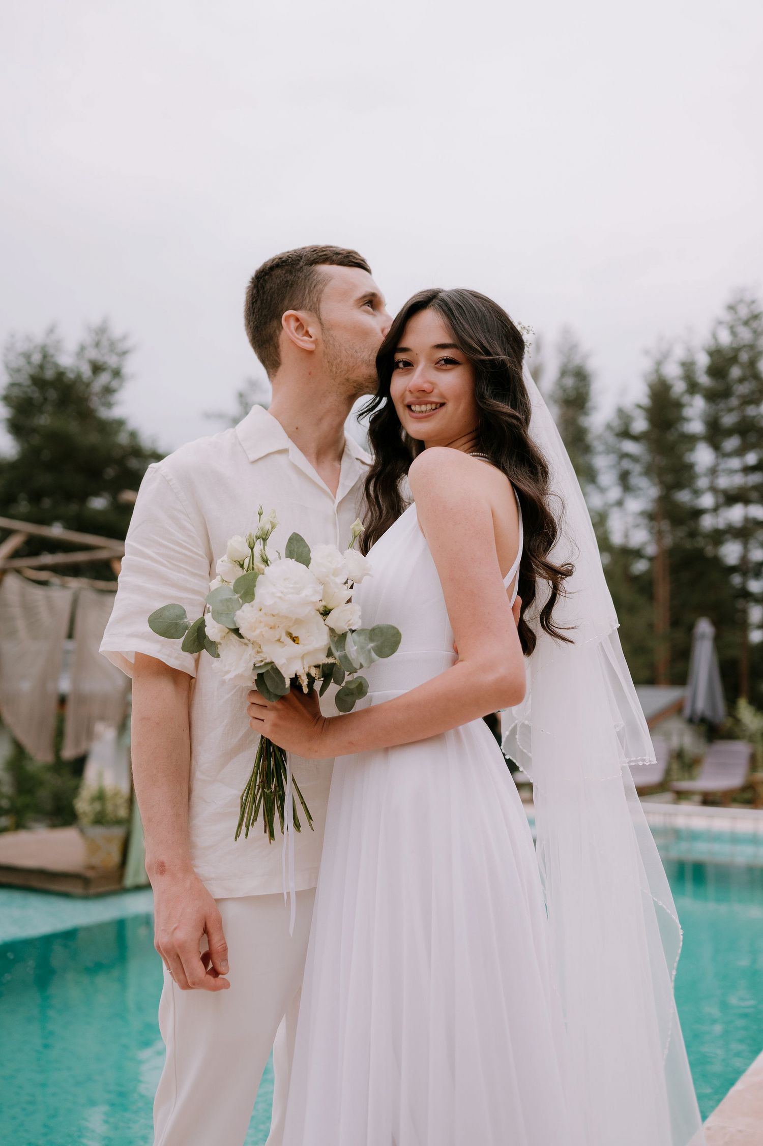 Newlyweds share joyful moments by a turquoise pool holding a white and green bouquet.