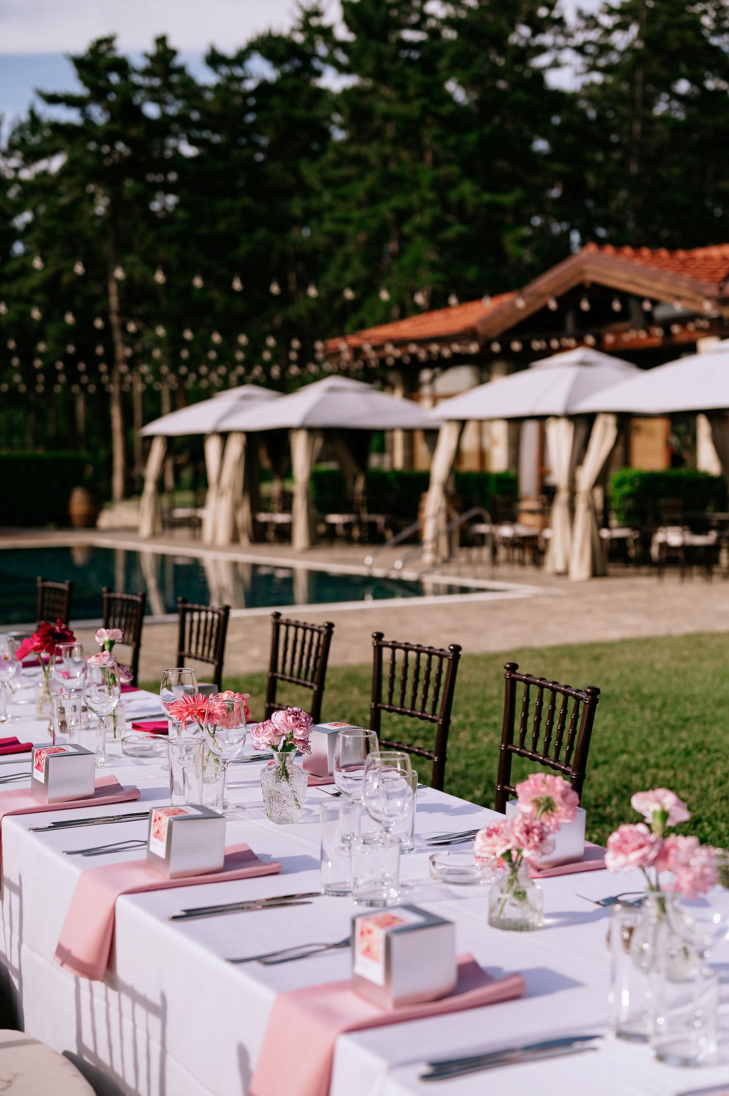 Romantic table setting with pink accents and string lights near a pool and covered cabanas.