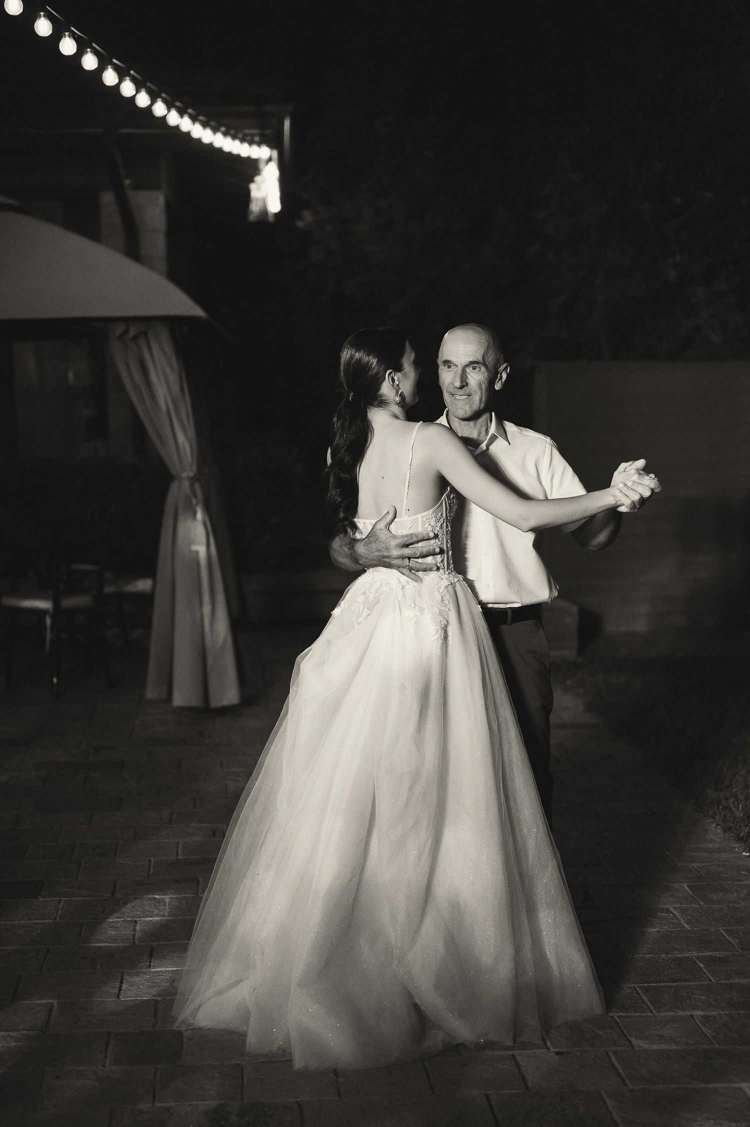 A couple dances beneath string lights on an outdoor patio during their wedding reception.
