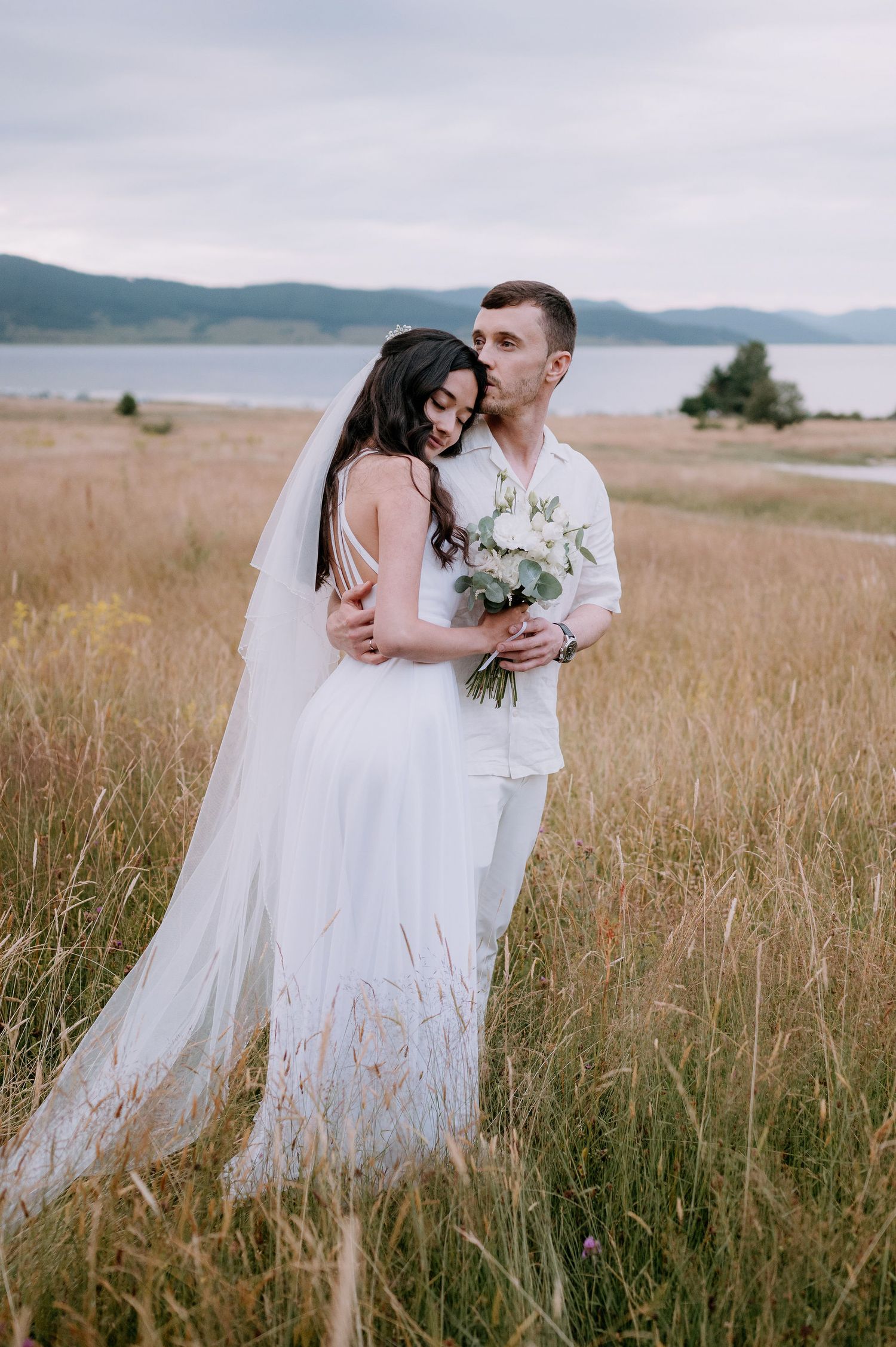 Bride in flowing dress and groom embrace while standing in tall grass near mountains.