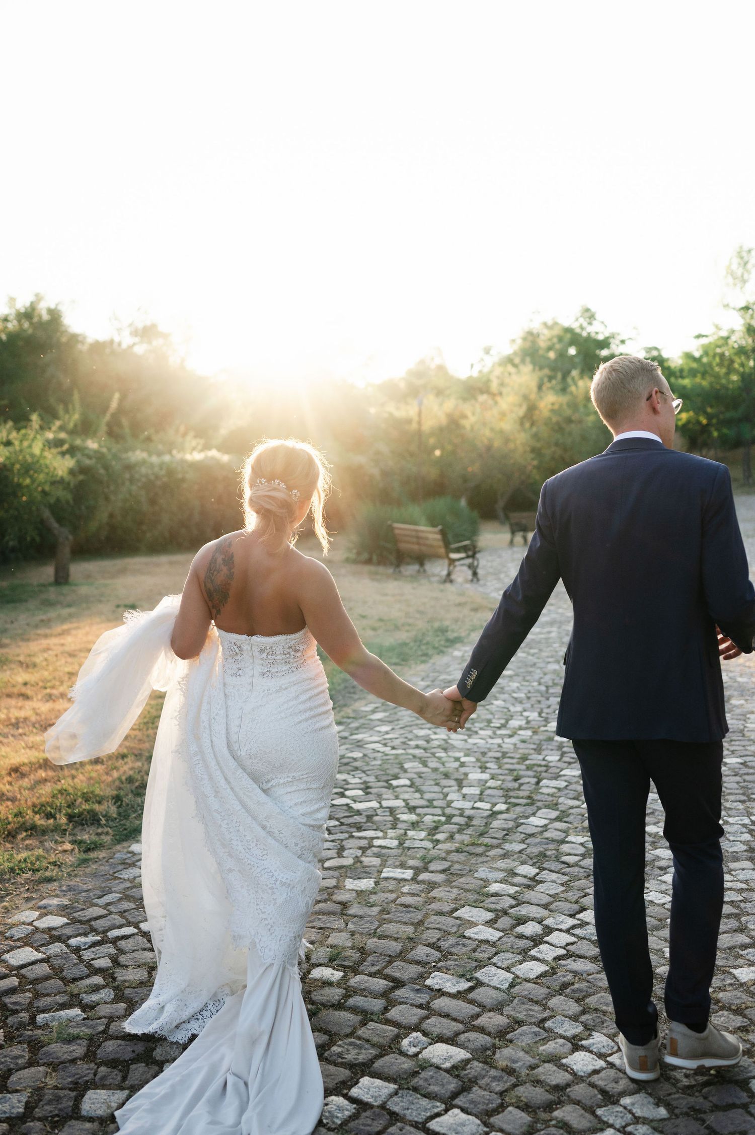 Bride in flowing white dress and groom walking hand in hand on a cobblestone path at dusk.