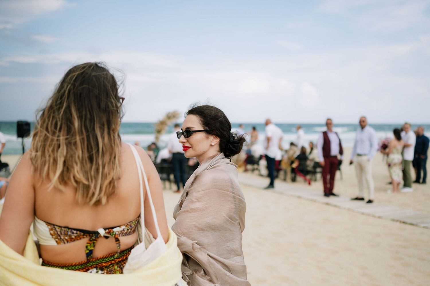 Wedding guests gather on a sunny beach with umbrellas and chairs set up for an outdoor ceremony.