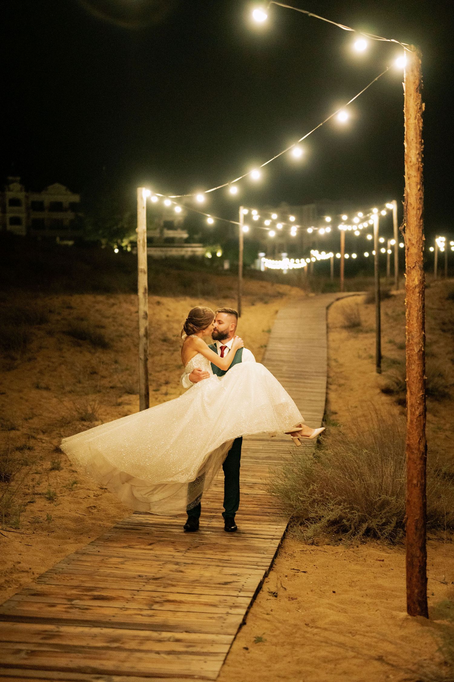 A couple in wedding attire dances romantically on a boardwalk under glowing string lights at night.