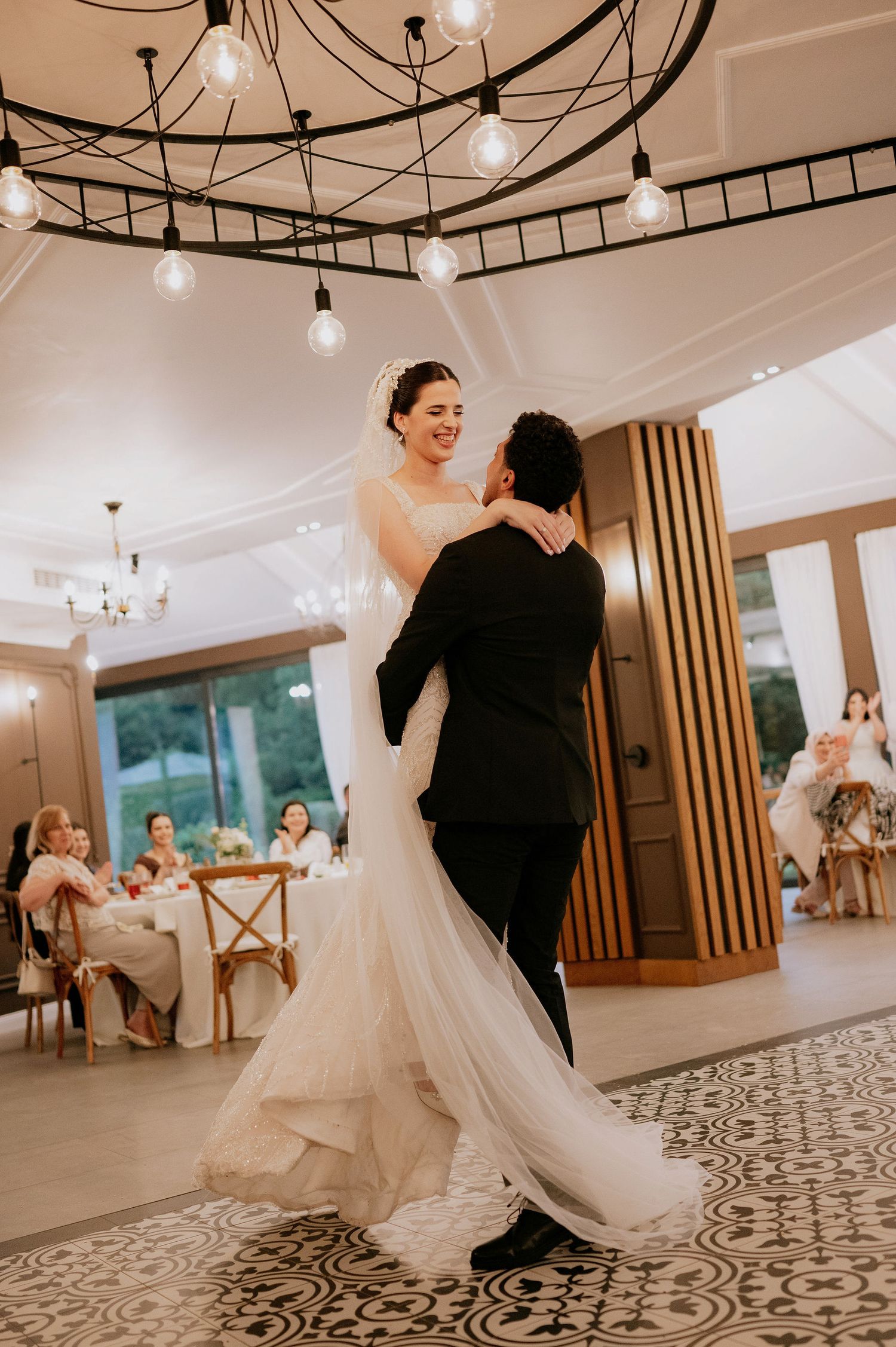 Wedding couple dances gracefully across a patterned ballroom floor under crystal chandeliers.