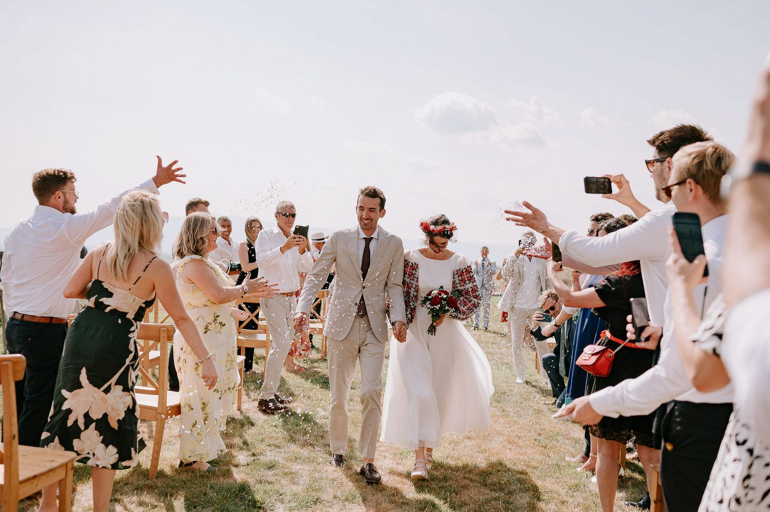 Wedding guests celebrate joyfully during an outdoor ceremony on a sunny hillside.