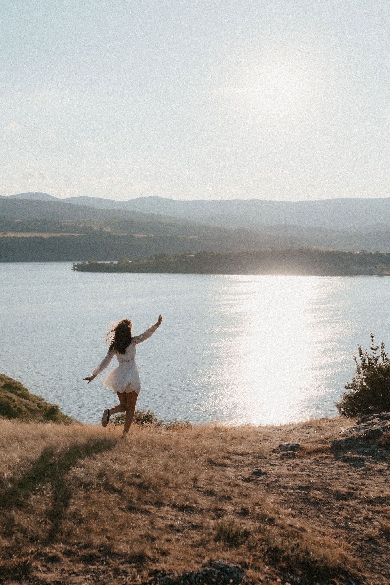 Person in white dress dancing freely on clifftop overlooking serene lake at sunset.