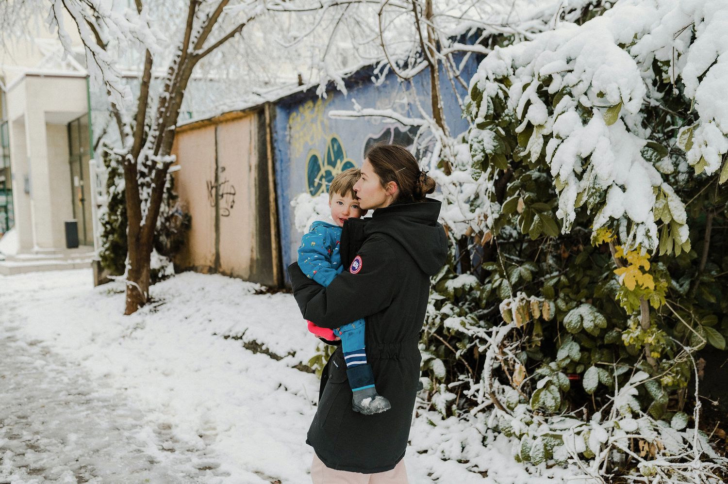 A person in winter attire takes photos in a snowy garden landscape with evergreen trees and bushes.