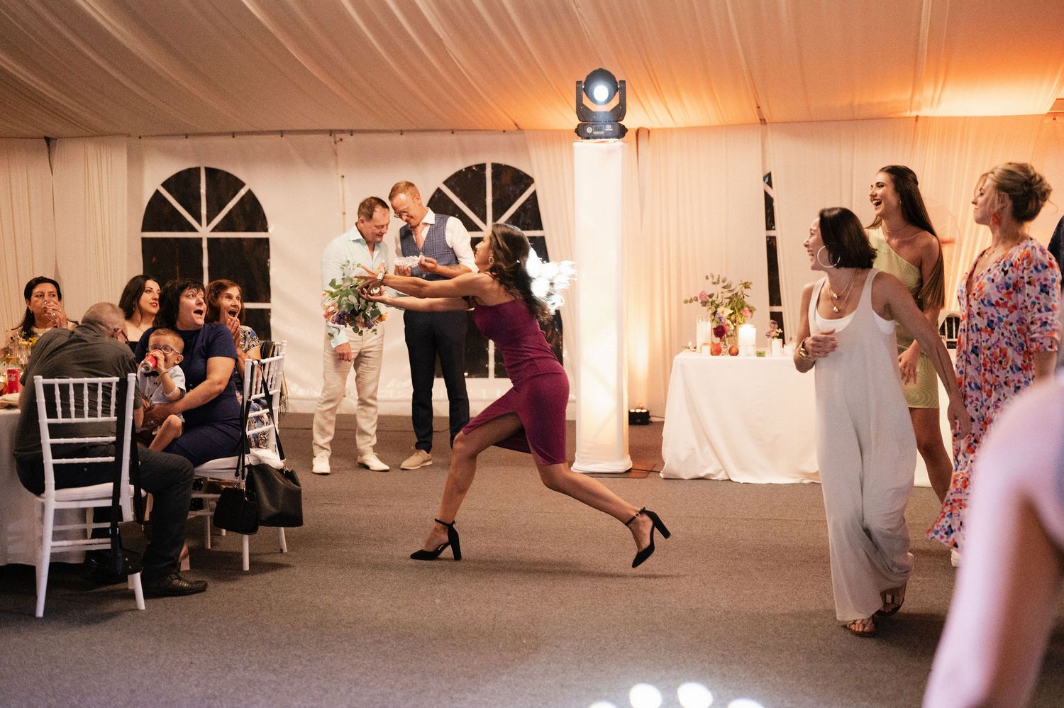 Wedding guests watch as two people perform an energetic dance move during a reception in a white tent venue at sunset.