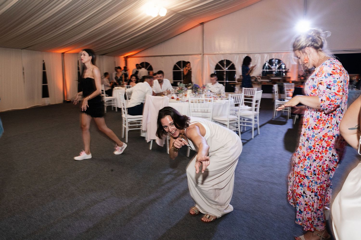 Wedding guests dance and bend down low on the dance floor inside a white tent reception venue with ambient lighting.