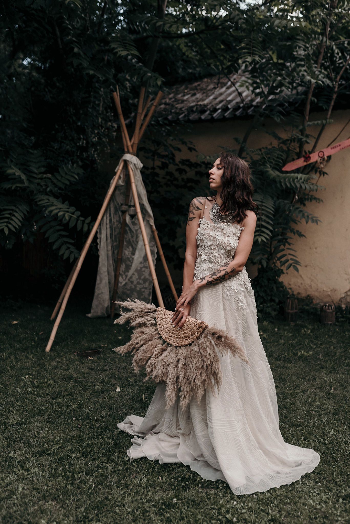A figure in a flowing white dress poses with decorative woven wall art in a garden setting.