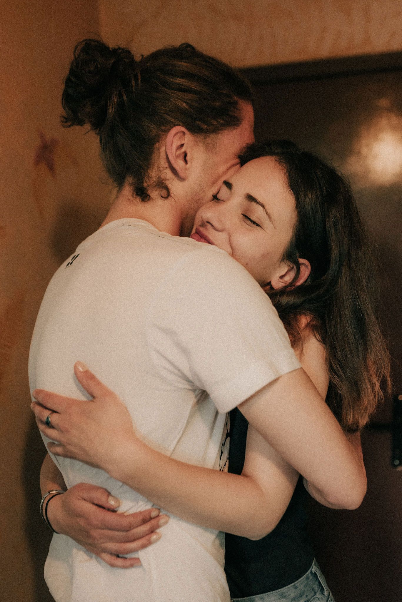 A couple in white and black clothing share an intimate embrace in a dimly lit room with warm lighting.