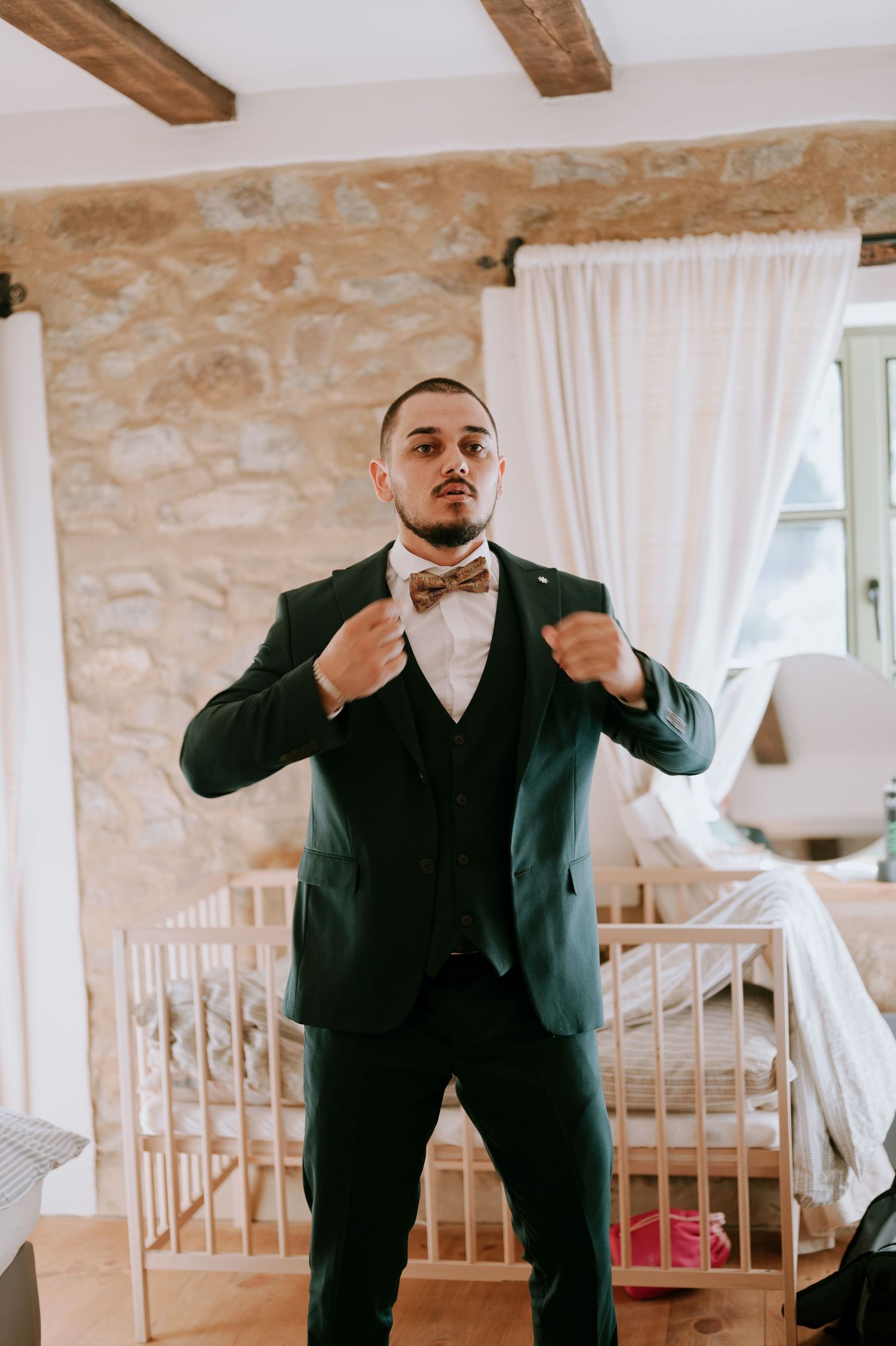 Someone in wedding attire adjusts their bowtie in a rustic room with stone walls.