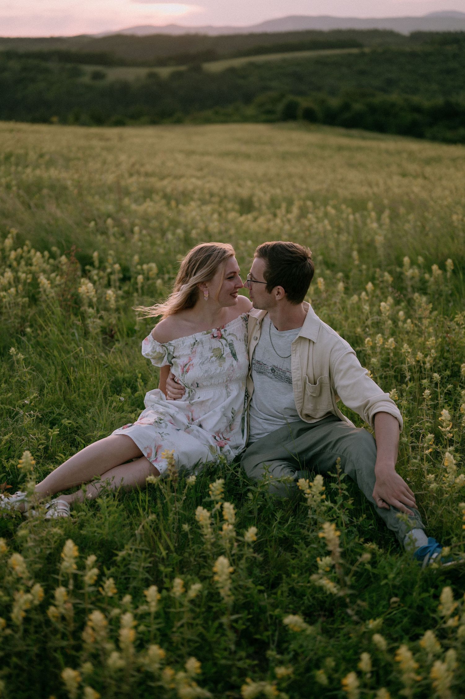 Romantic couple sits together in a blooming wildflower meadow at sunset with rolling hills in the background.