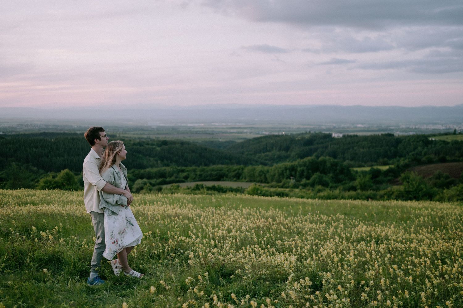Two people stand together in a golden field overlooking a vast landscape during dusk.