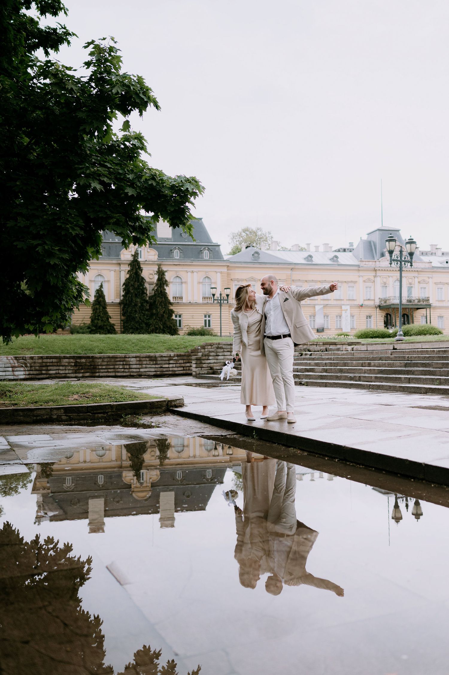 Couple in light colored outfits dancing near reflecting pool with historic palace building in background.