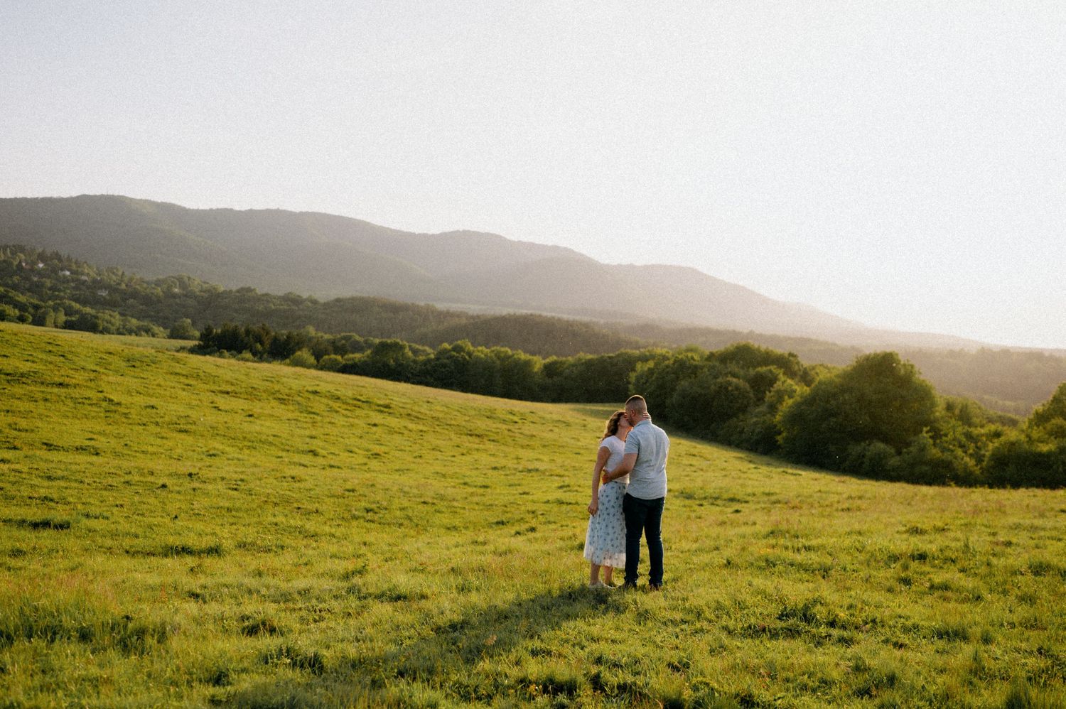 A panoramic view of a couple dancing in a vast green field with mountains in the distance.