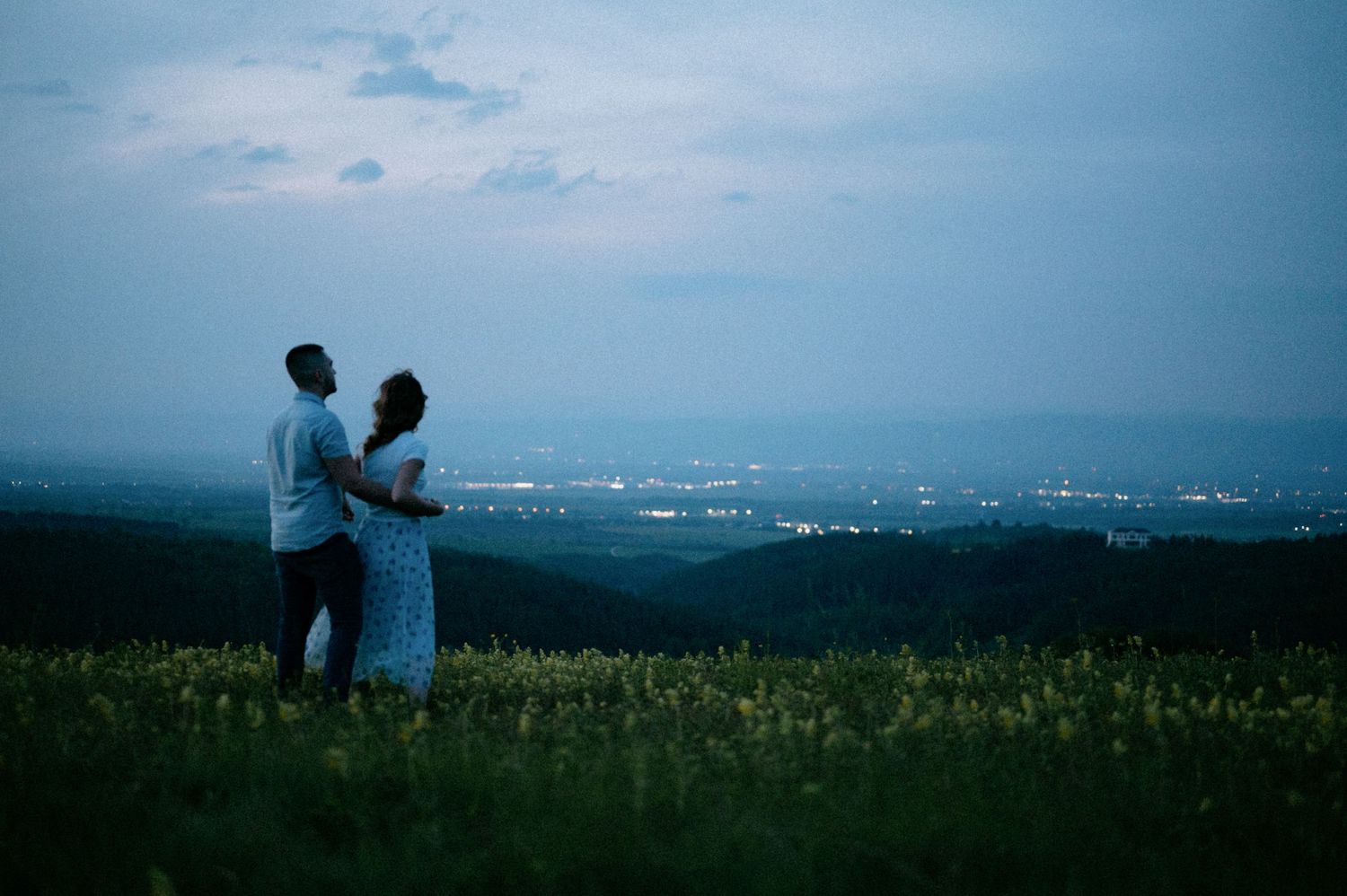 Two figures embrace on a hilltop with a panoramic view of city lights below.