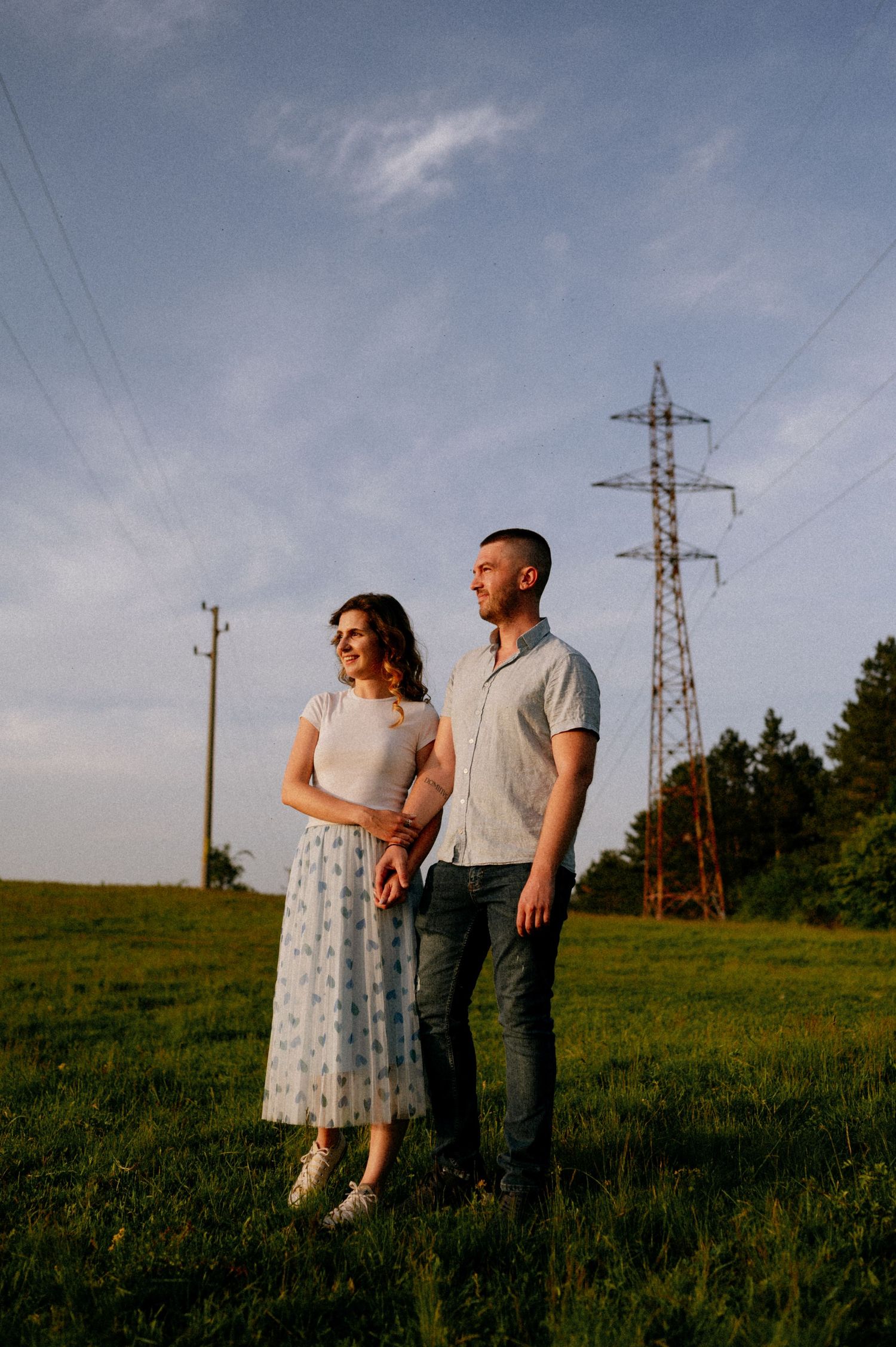 A romantic couple poses in a field at sunset with electrical towers in the distance.