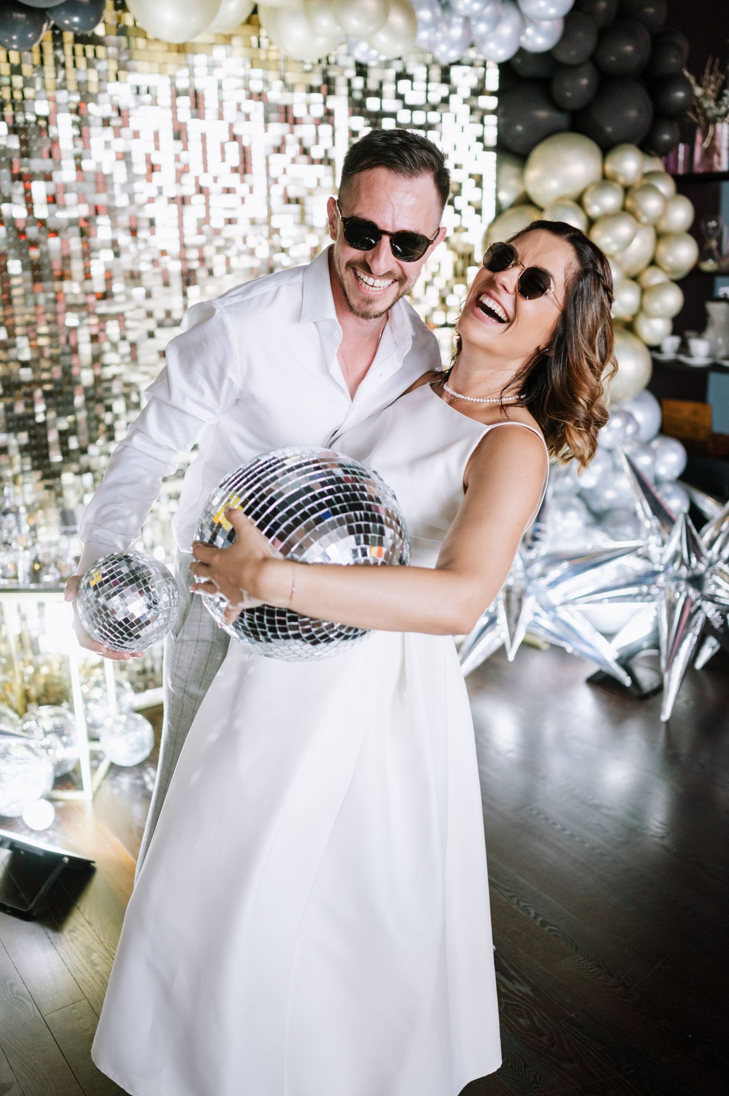 Couple in white outfits laughing while holding disco ball against sparkly backdrop with balloon decorations.