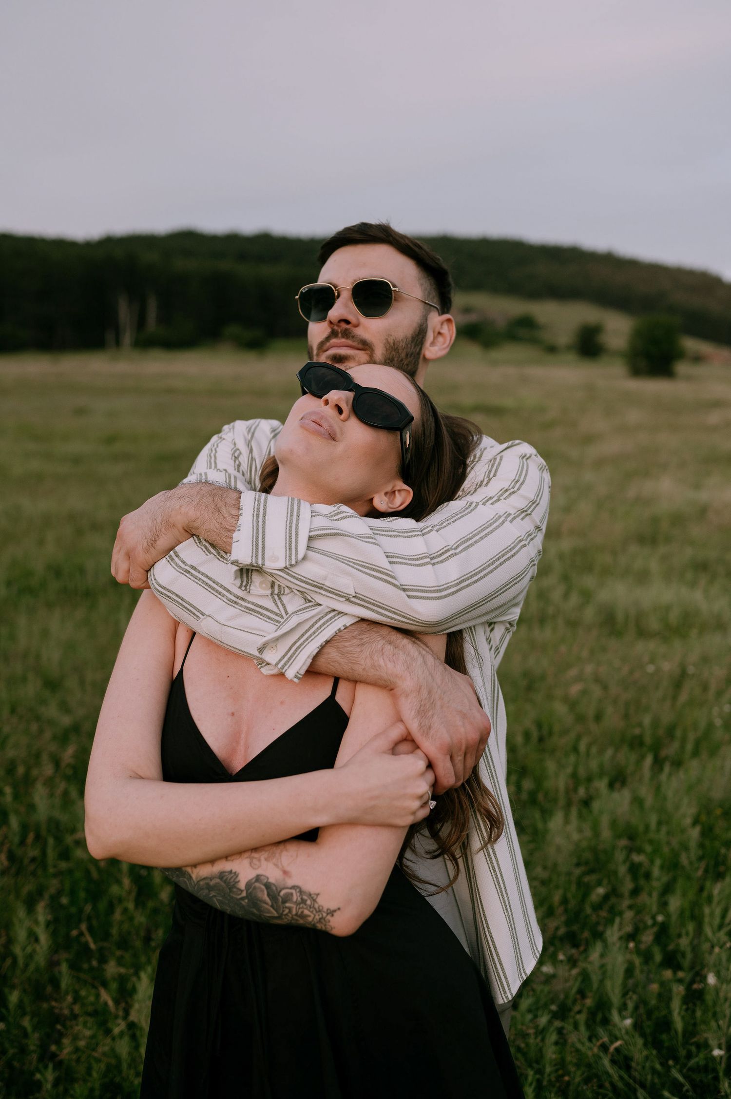 Two people embracing in a grassy field while wearing sunglasses and casual attire.