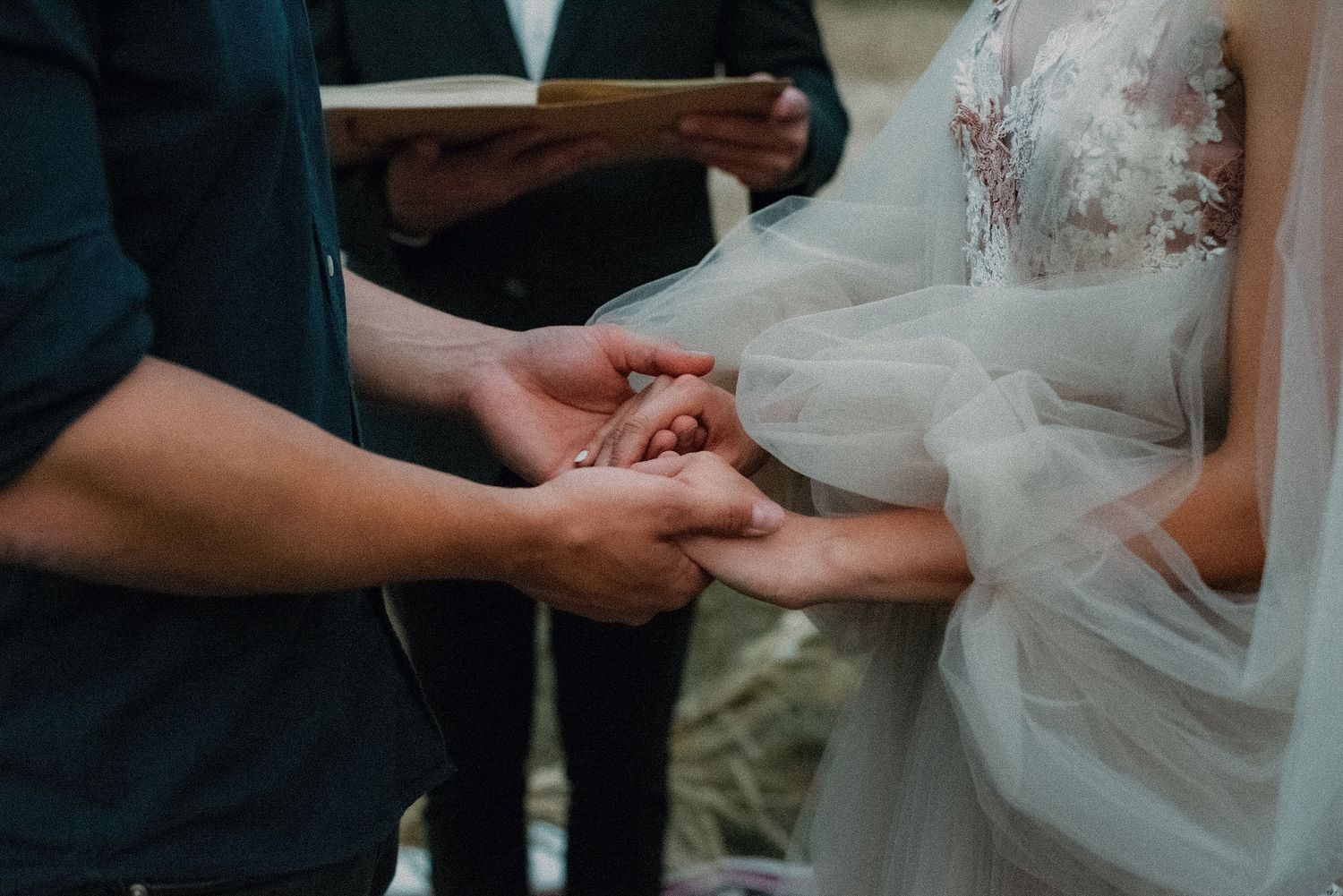 Close-up of hands exchanging rings during an outdoor wedding ceremony with a lace wedding dress visible.