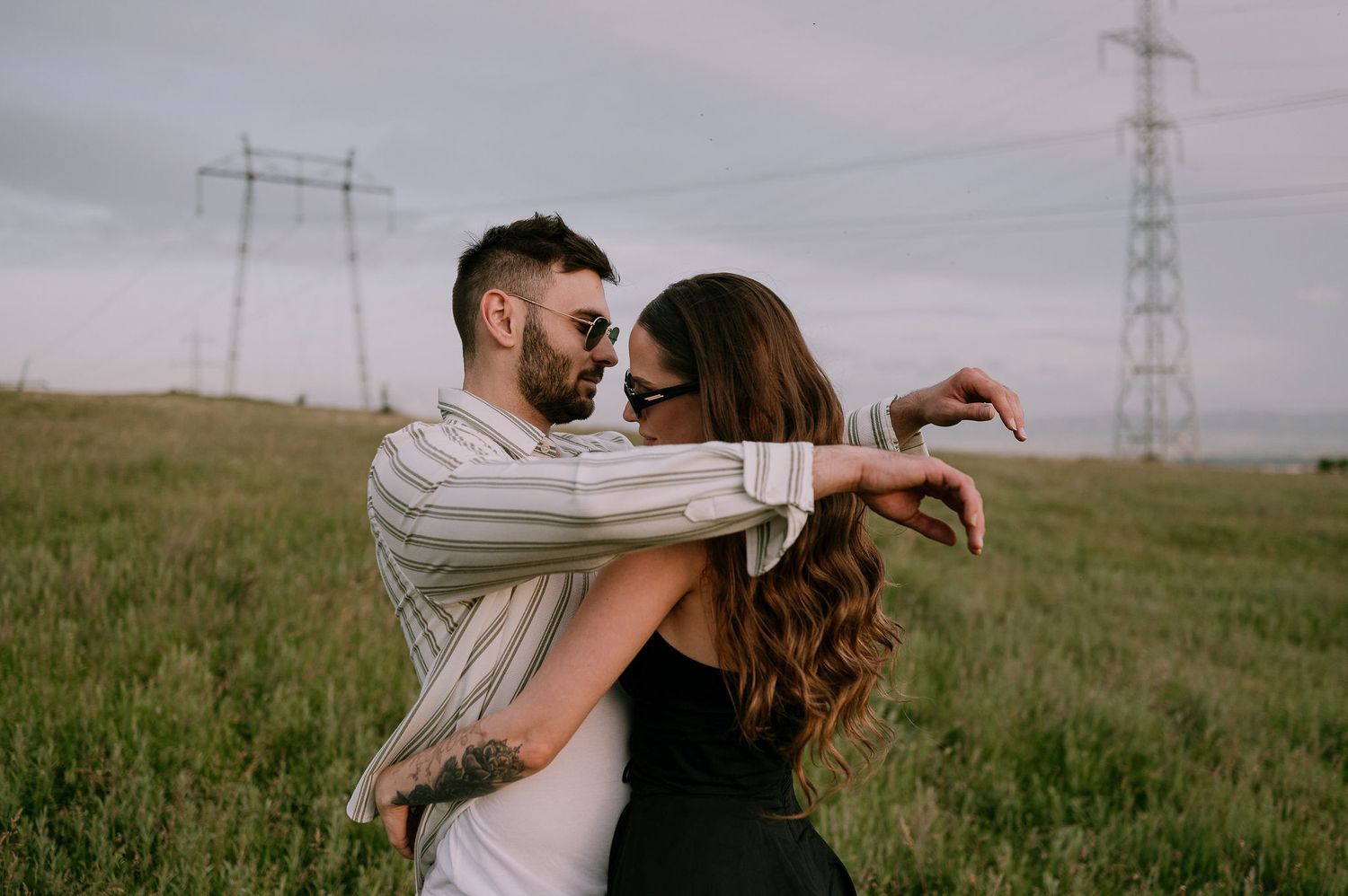 A couple embraces in a grassy field with electrical transmission towers in the background under an overcast sky.