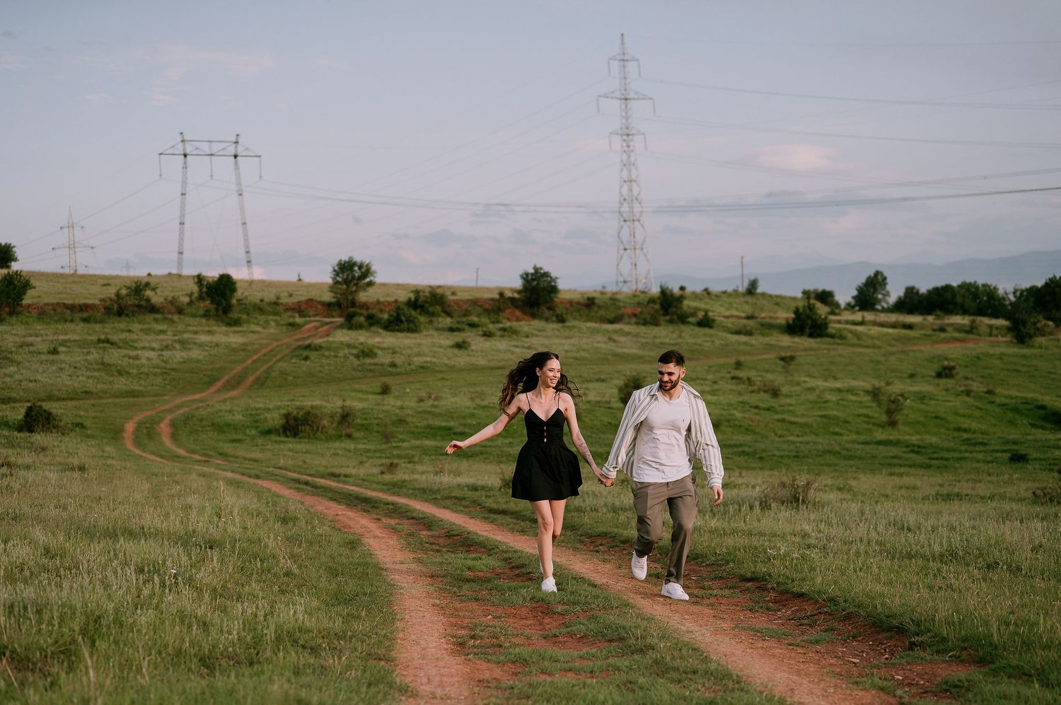 A couple walks hand in hand along a winding dirt path through a green rural landscape with power lines in the distance.