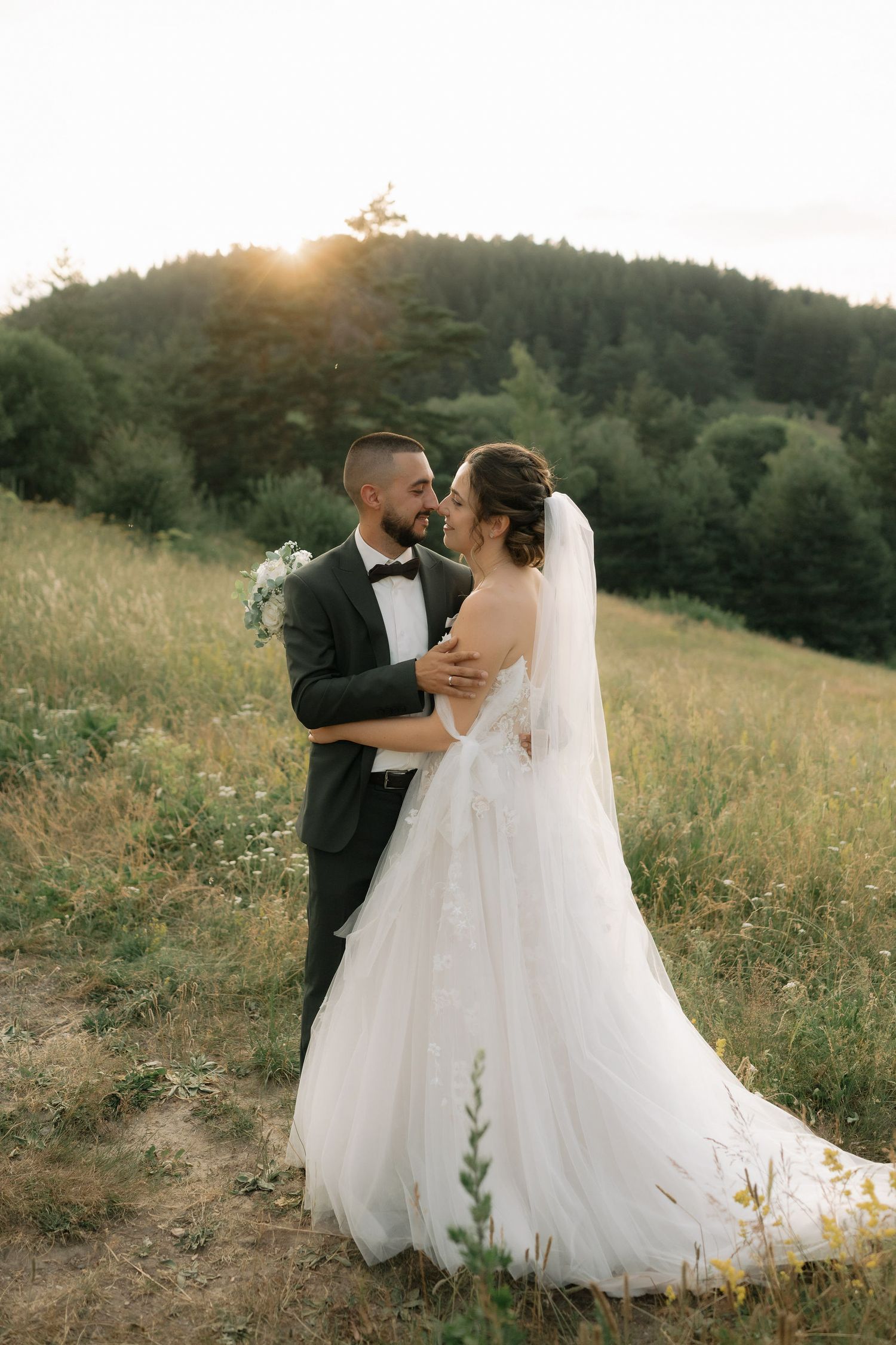 A couple embraces in a romantic sunset moment on a grassy hillside, with the bride wearing a flowing white gown and veil.