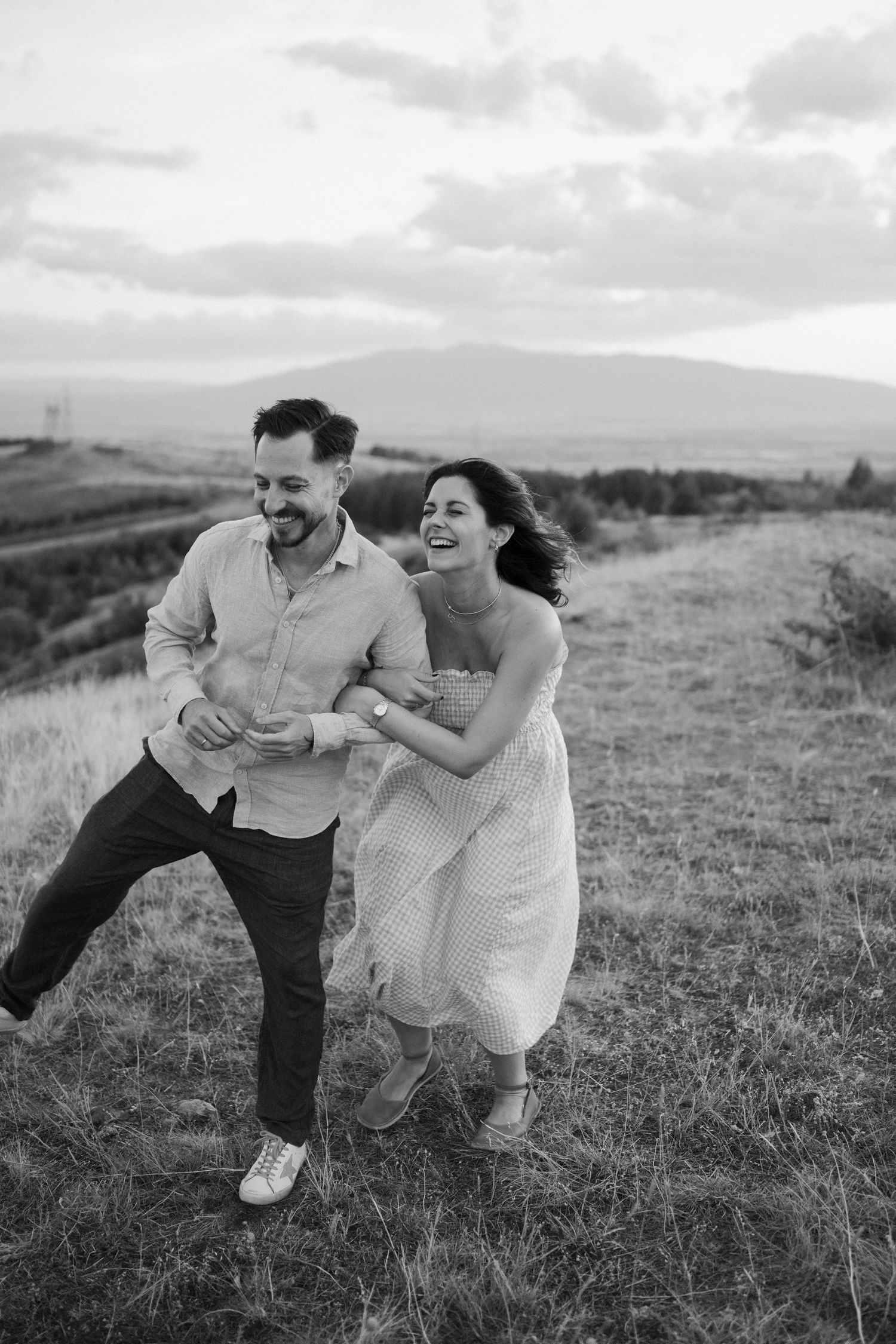 A couple shares joyful laughter while running together on a grassy hillside in black and white photography.