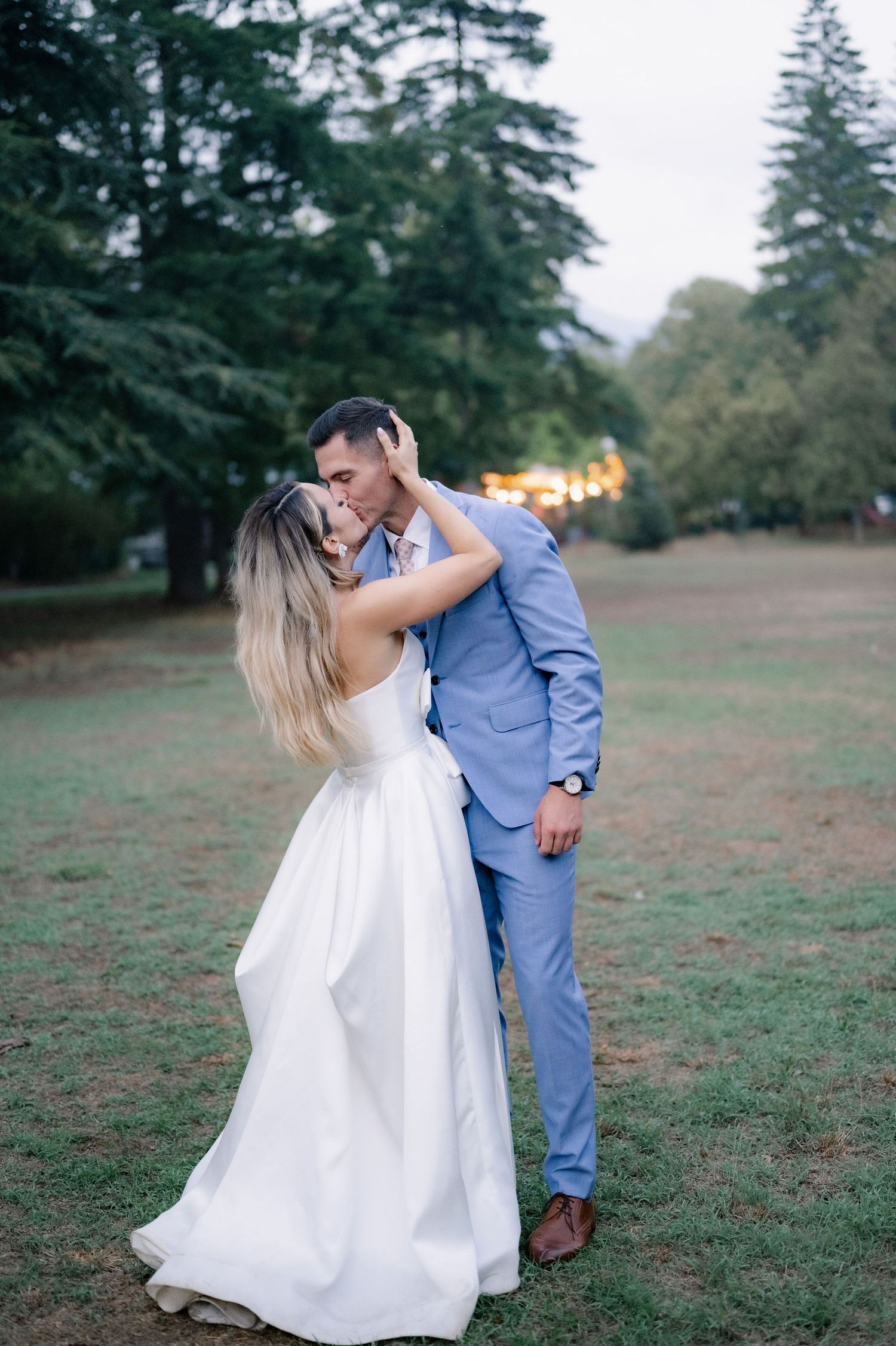 A couple in wedding attire dance together in a grassy field surrounded by tall pine trees at dusk.