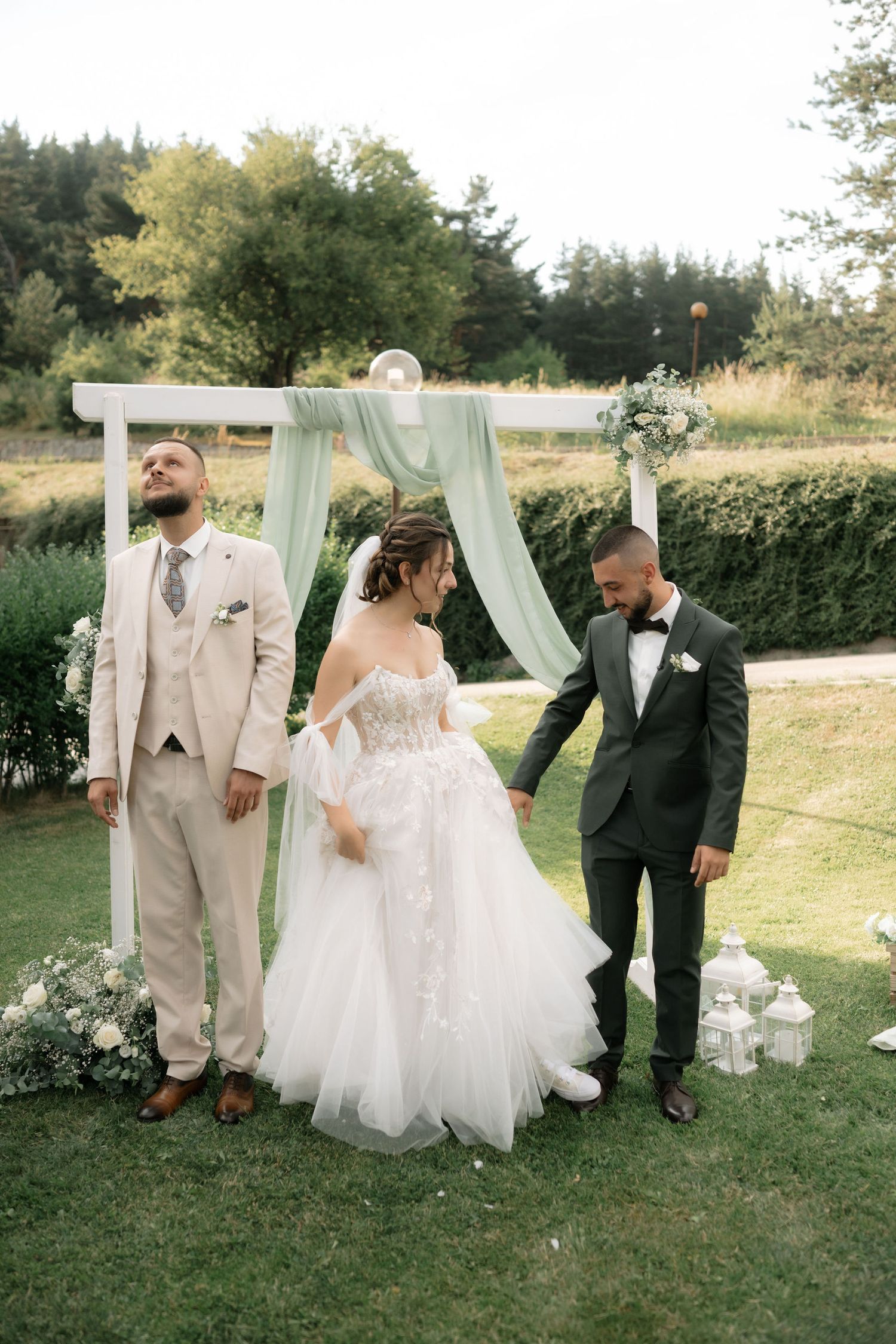 Wedding party members pose together on a grassy lawn with decorative white curtains and lanterns.