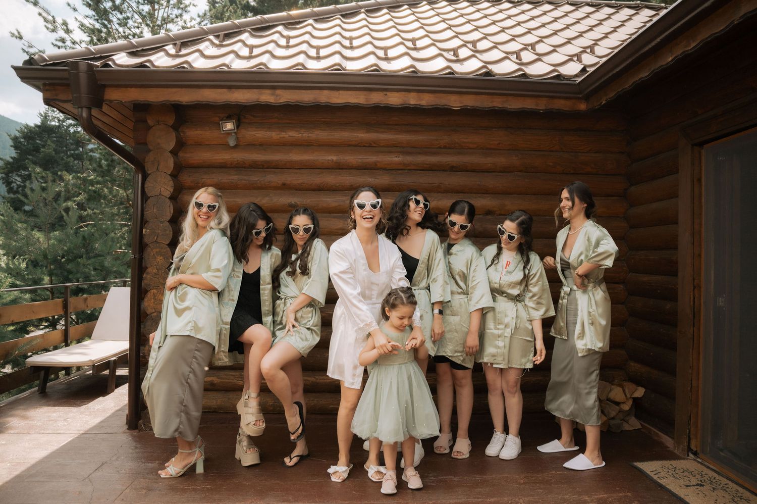 Wedding group stands in coordinated beige and white outfits against wooden lodge exterior.