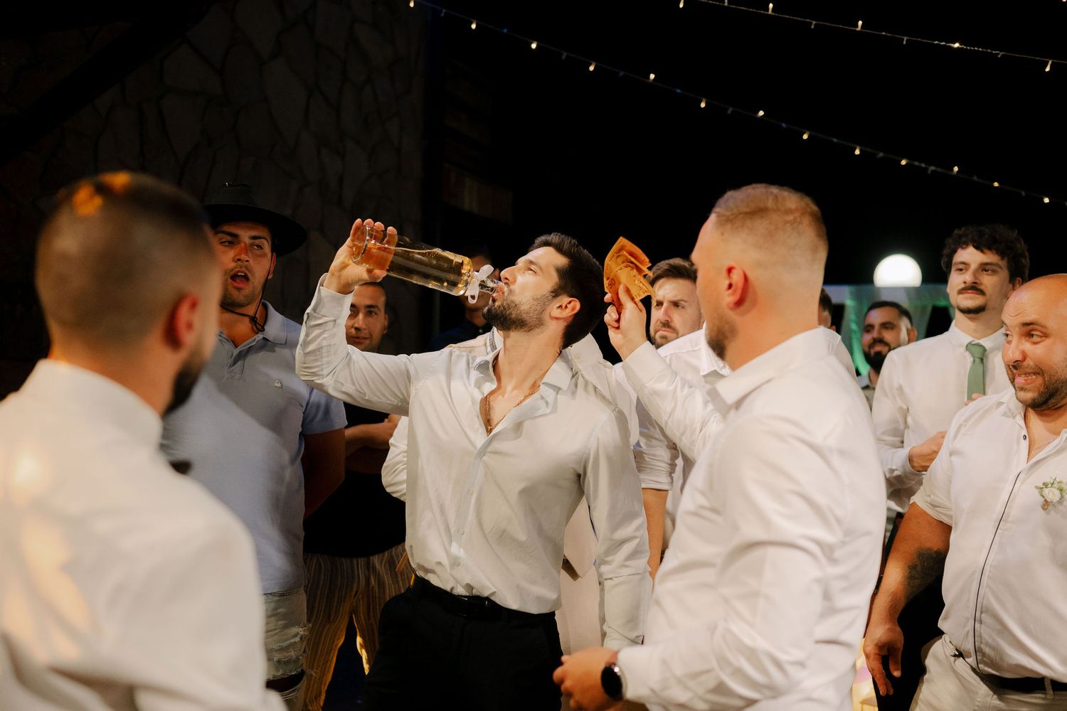 A group of men in white shirts link arms while drinking together in a celebratory toast at a gathering.