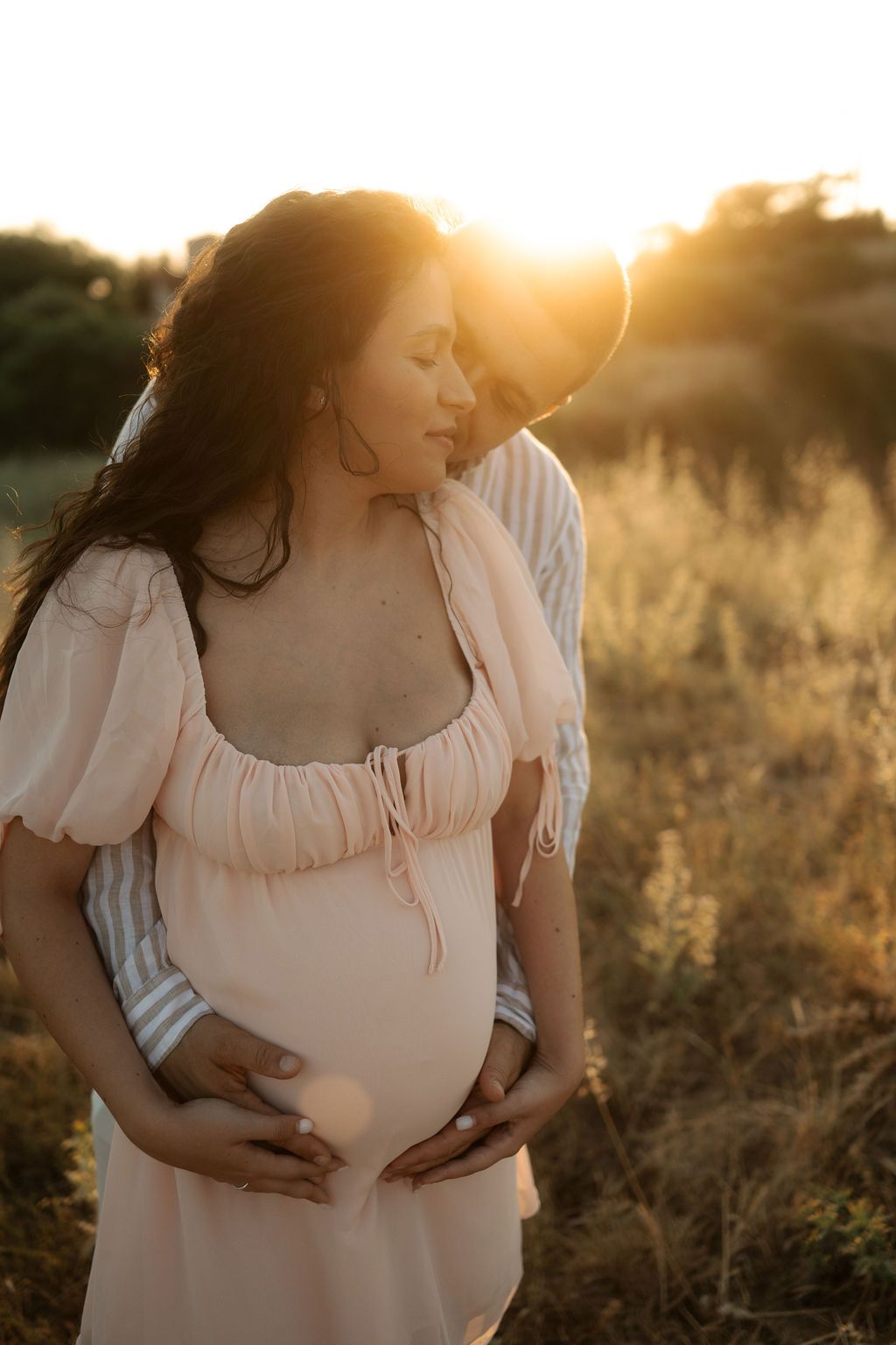 Maternity photoshoot in a golden field at sunset shows a flowing white dress and gentle hands cradling a baby bump.