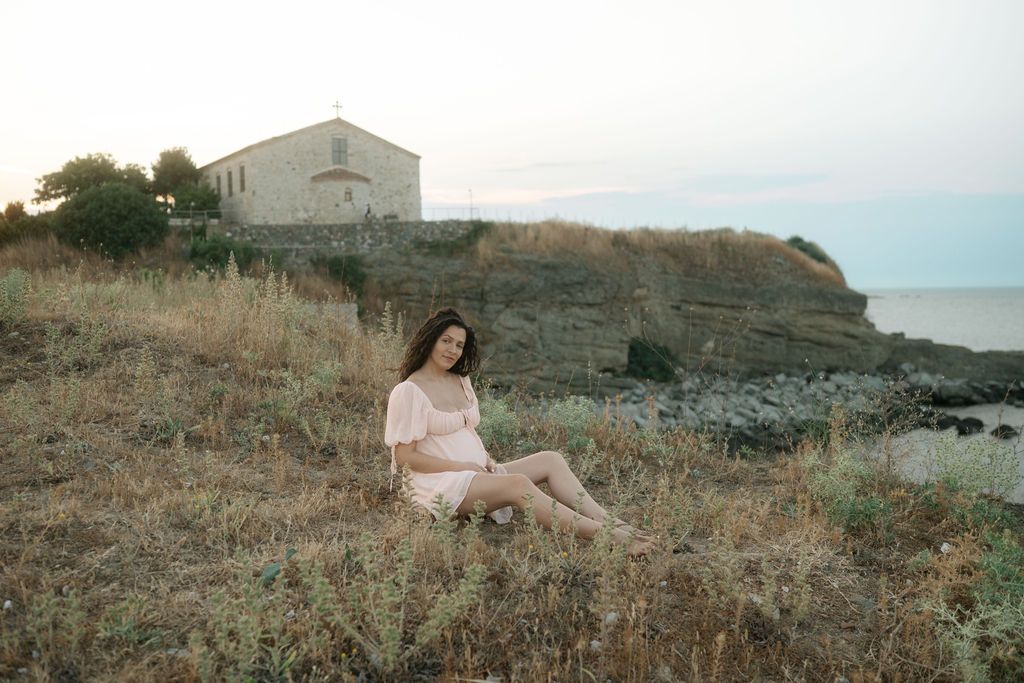 A person in a pink dress sits on a grassy hillside overlooking the ocean at sunset.