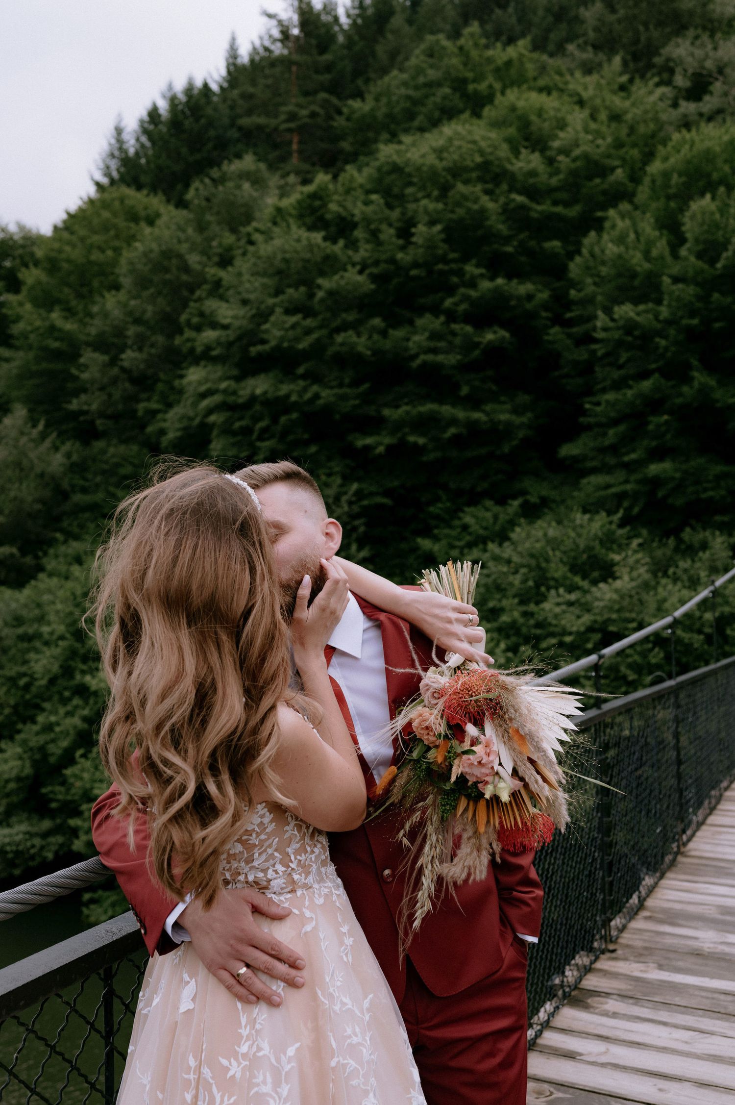 A couple shares romantic kisses while walking across a wooden suspension bridge surrounded by lush forest.