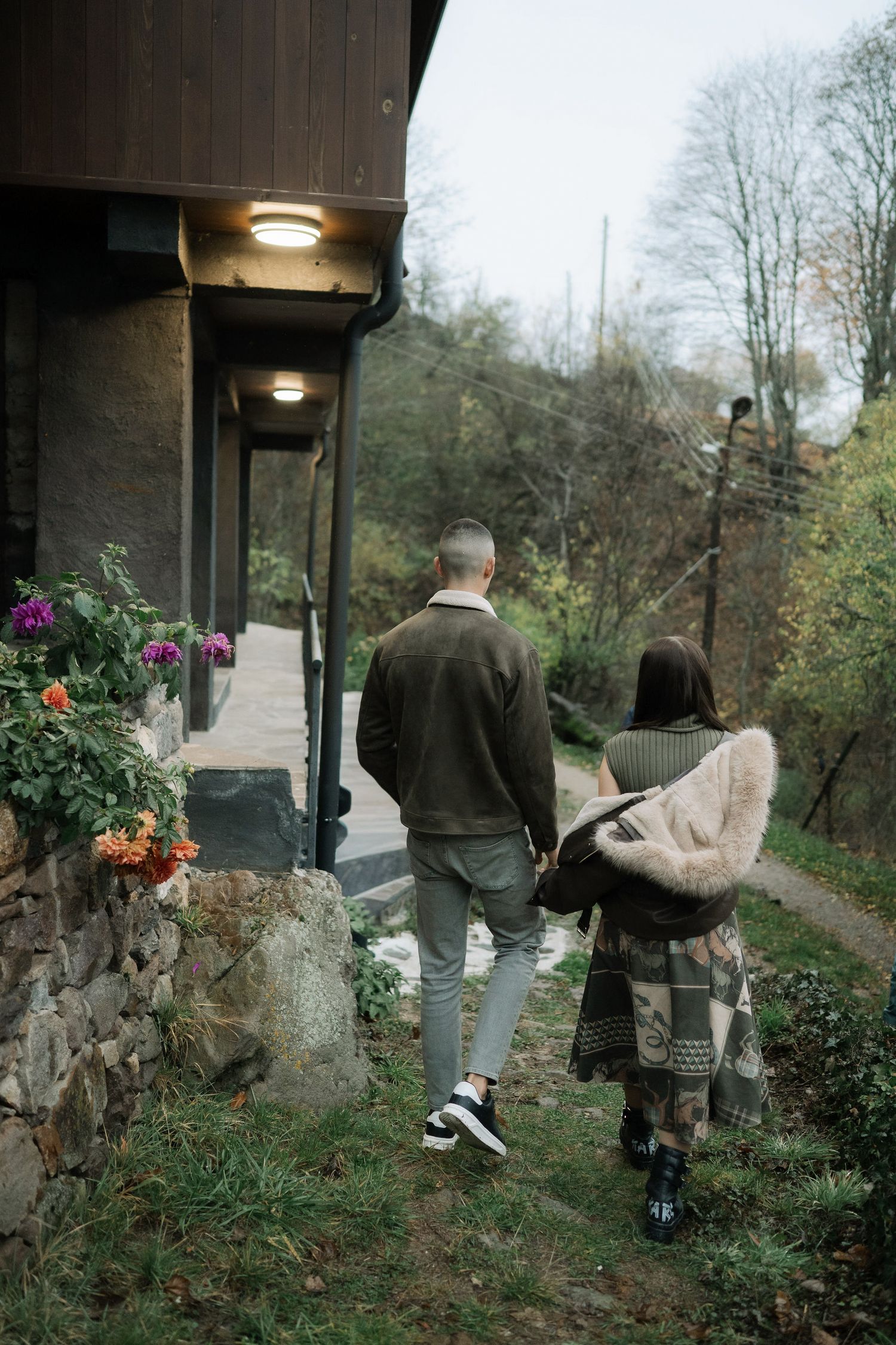 People walking along a stone wall path surrounded by greenery and flowers in a garden setting.