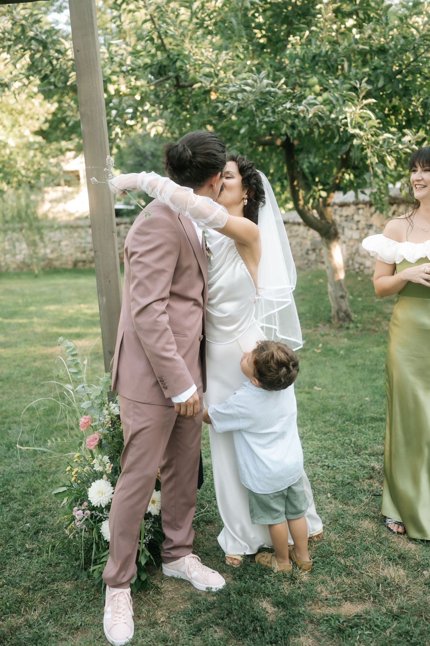 A romantic wedding couple shares a playful dip kiss in a garden setting with green trees in the background.