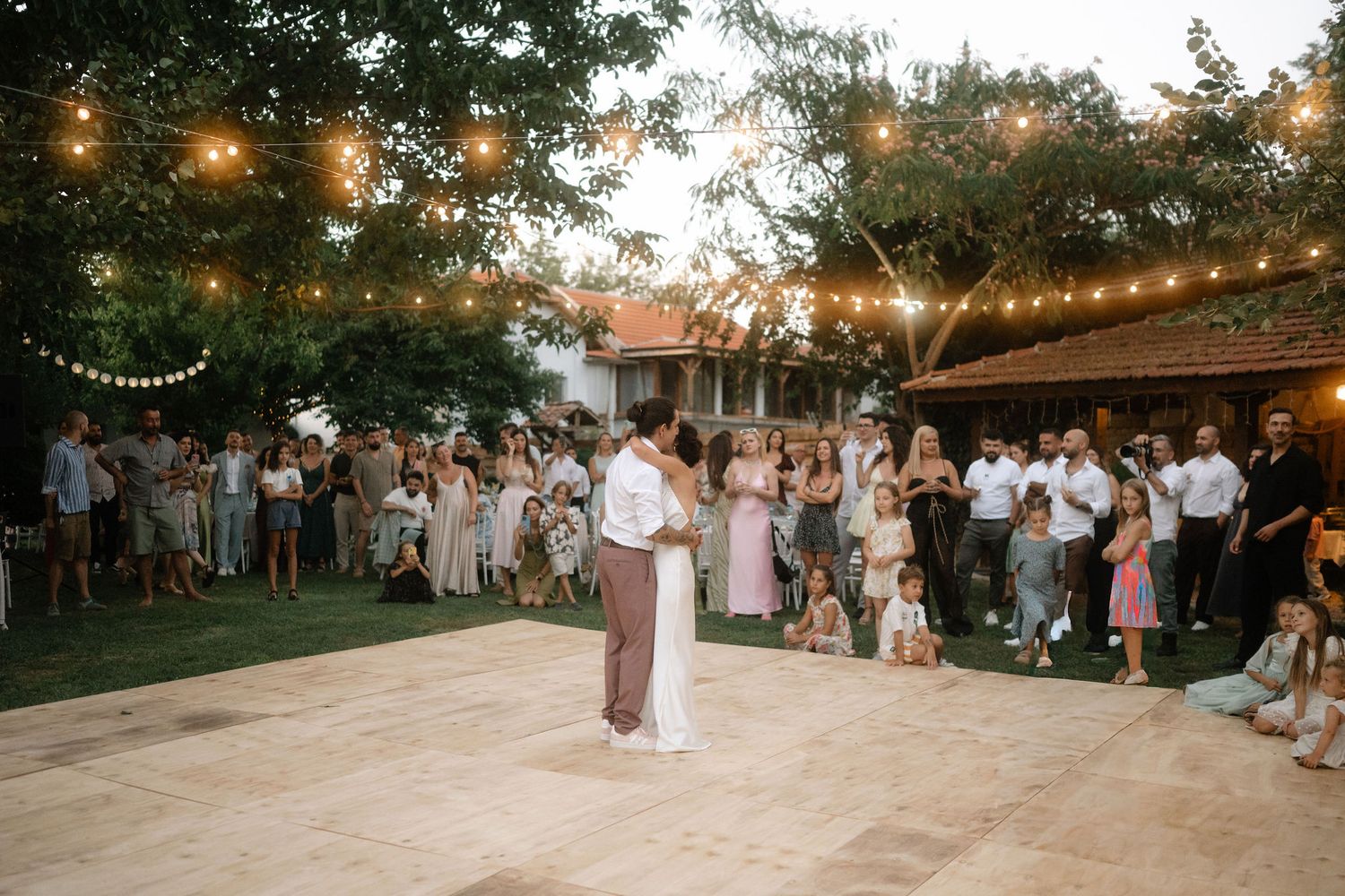 A string of warm glowing lights illuminates an outdoor dance floor as people gather for an evening celebration.