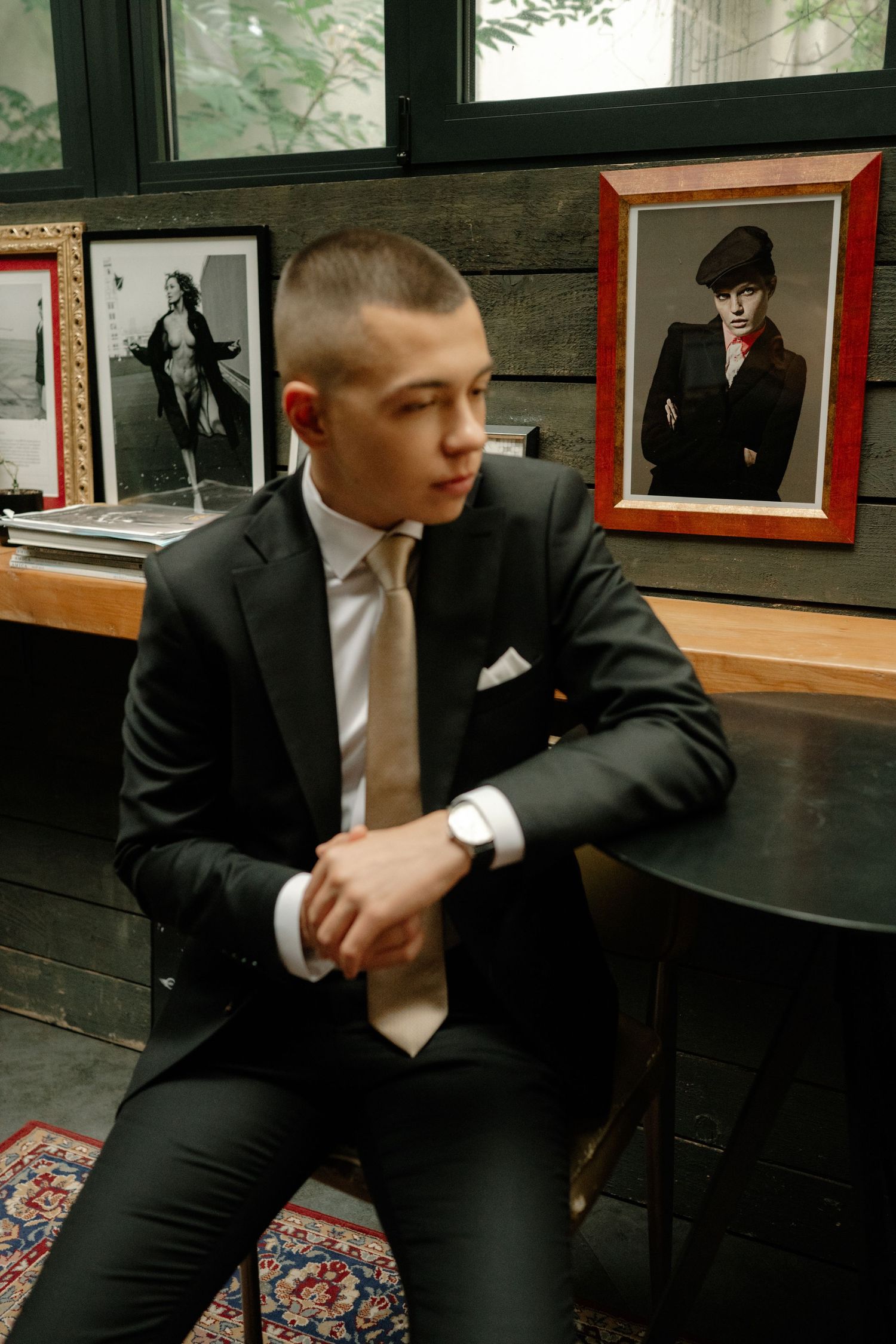 A person in a black suit sits on a patterned carpet in what appears to be a formal setting.
