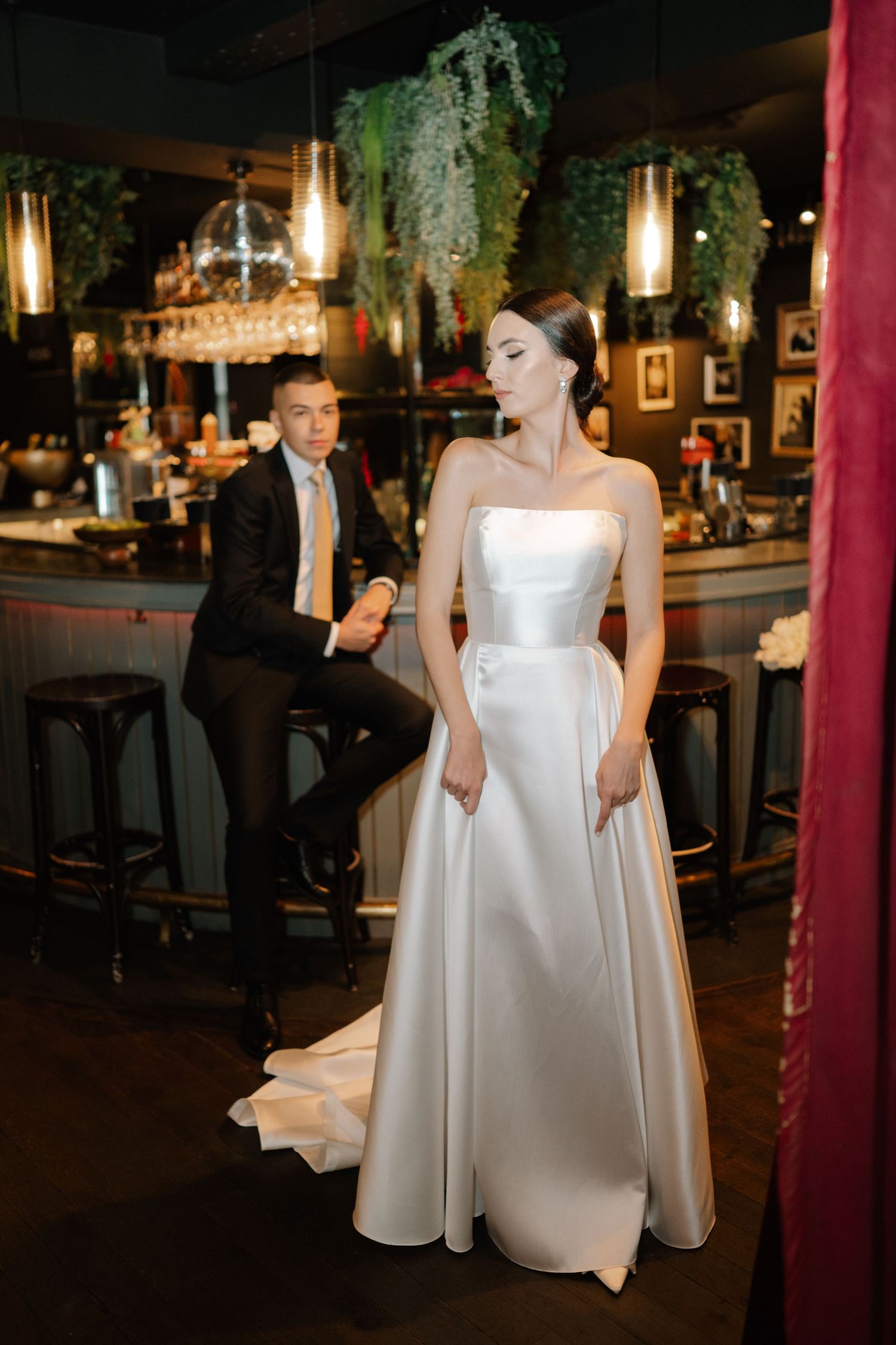 A bride in a modern white satin dress twirls on a dark wooden dance floor under pendant lighting at an indoor venue.