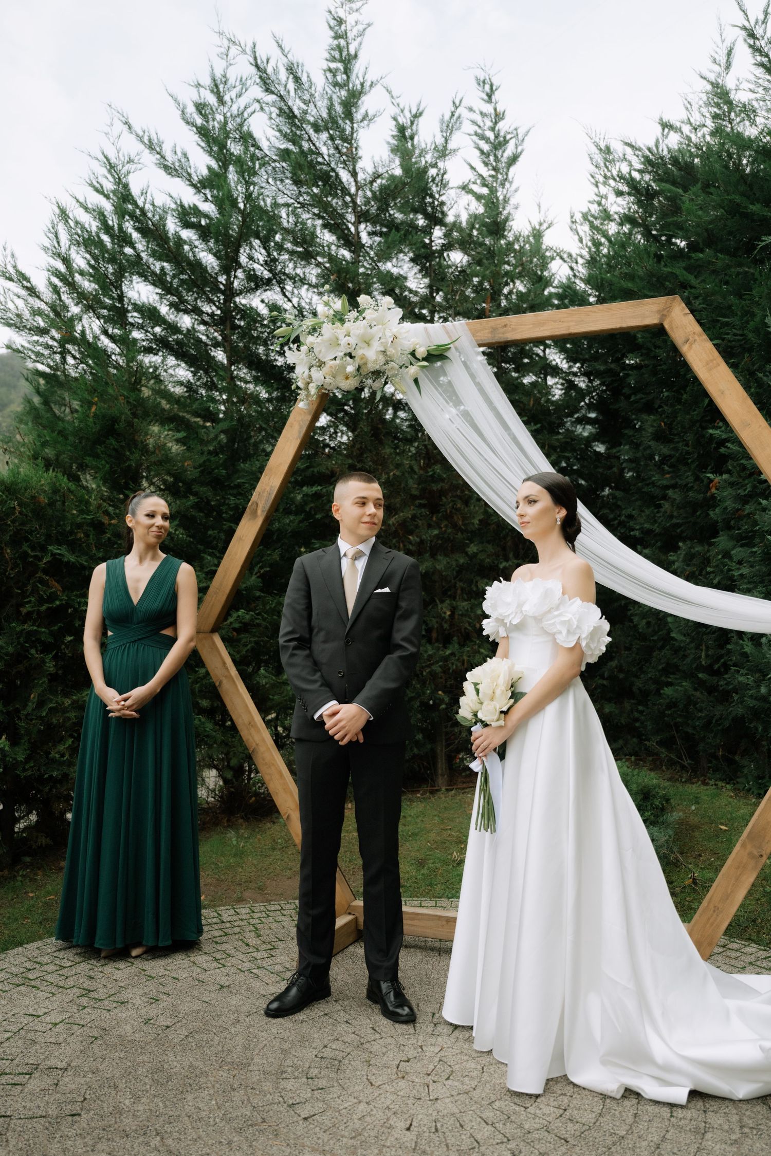 An outdoor wedding ceremony setup with a wooden hexagonal arch decorated with white fabric and florals against evergreen trees.