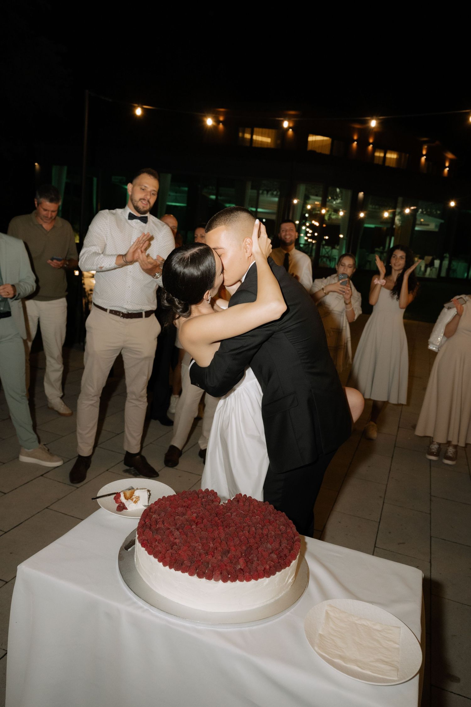 A couple shares a romantic kiss behind their red-topped wedding cake at an evening reception while guests look on.