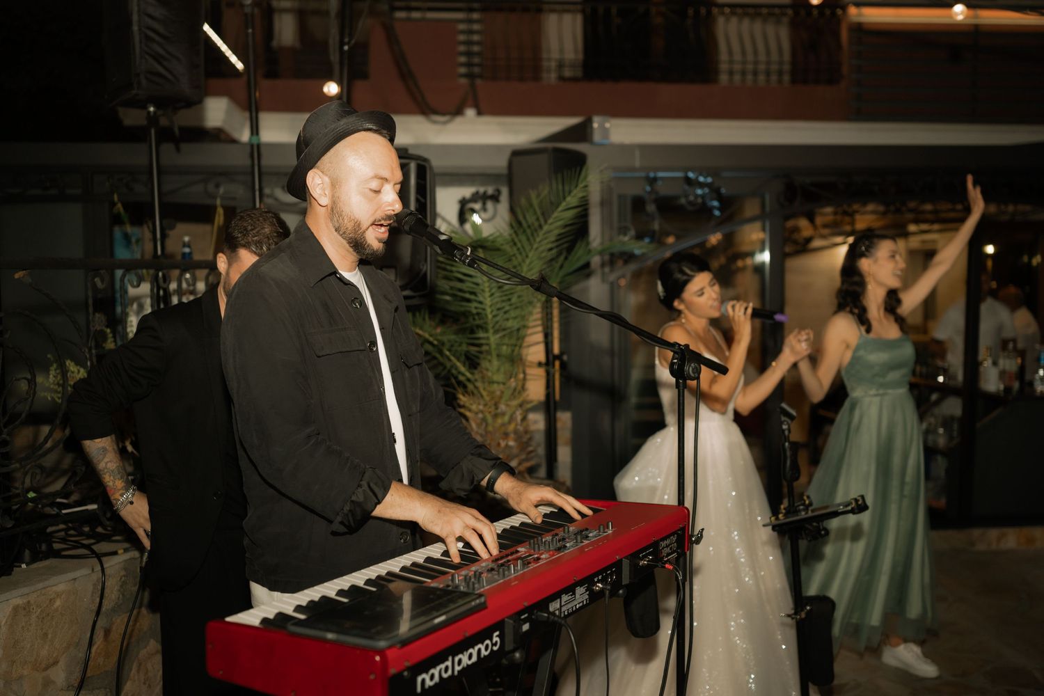 Musician plays a red Nord keyboard at an evening event while performers sing and dance on stage.