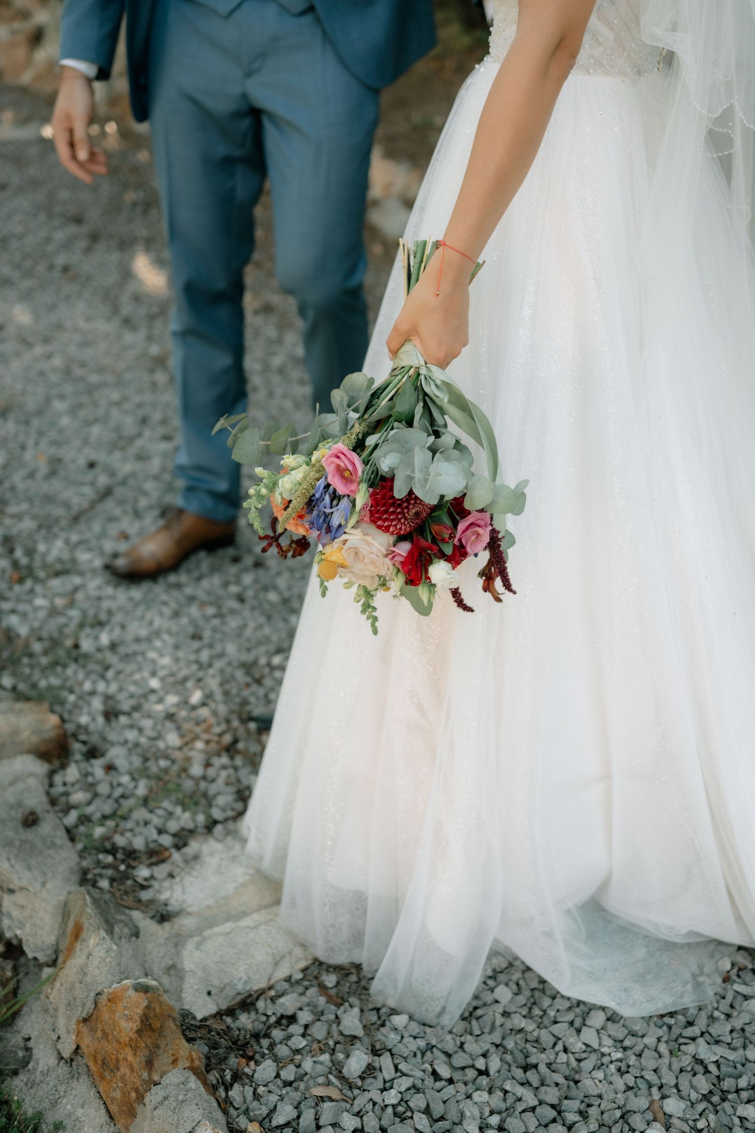 Close-up series of wedding details showing white tulle dress and burgundy floral bouquet against gravel ground.