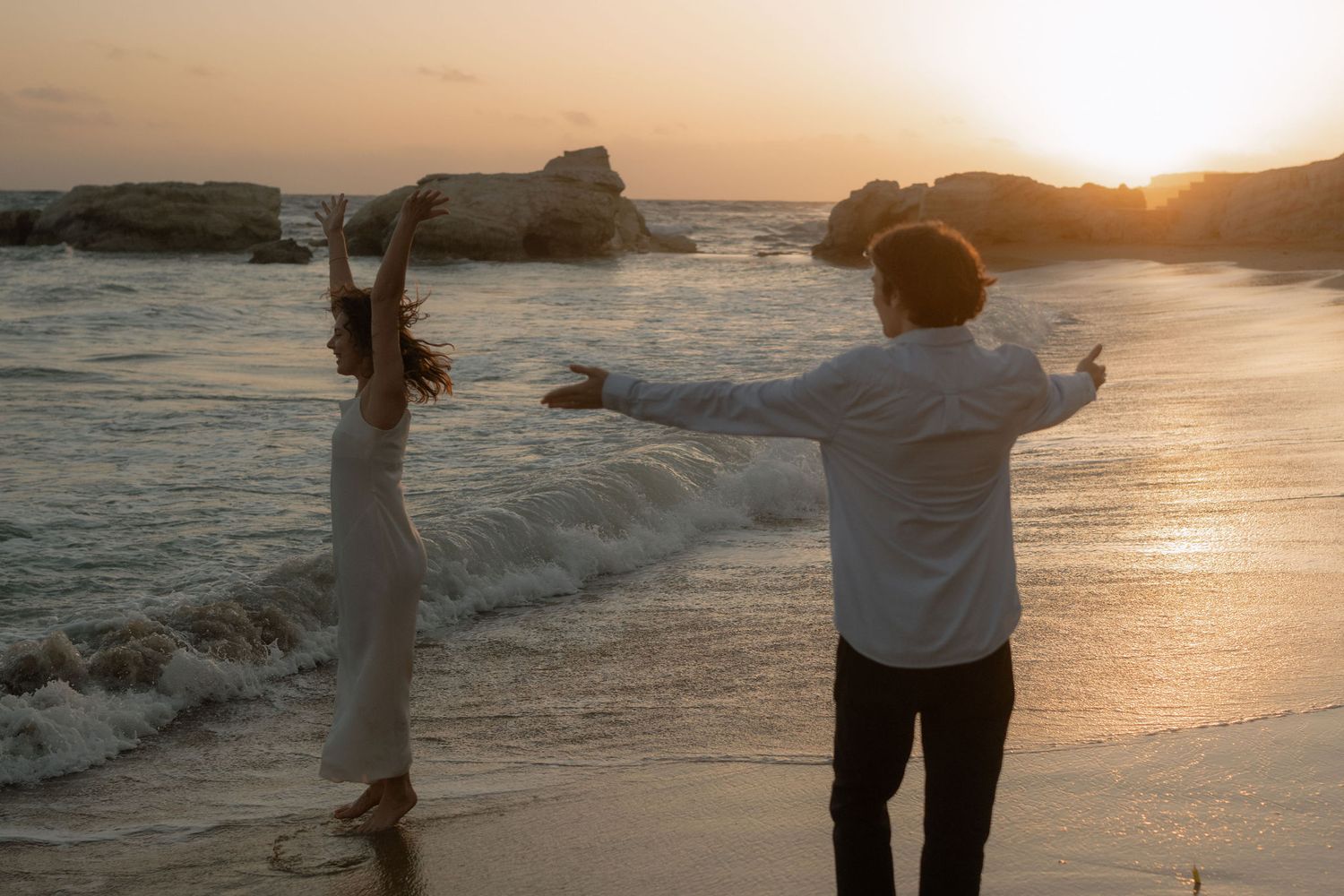 Couple dancing romantically on a sandy beach at golden sunset with ocean waves and rocks in background.