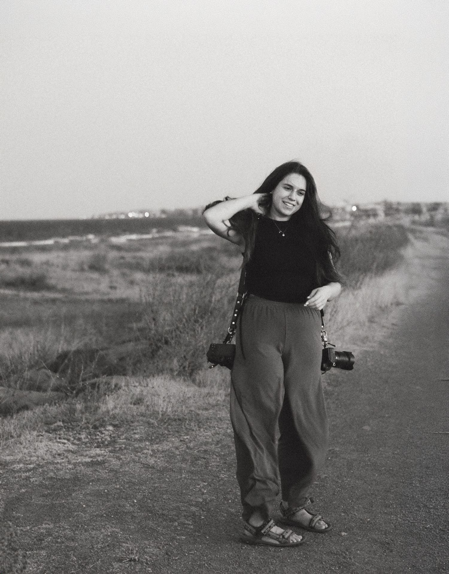 Woman smiling on coastal path holding cameras, black and white photography portrait outdoors.