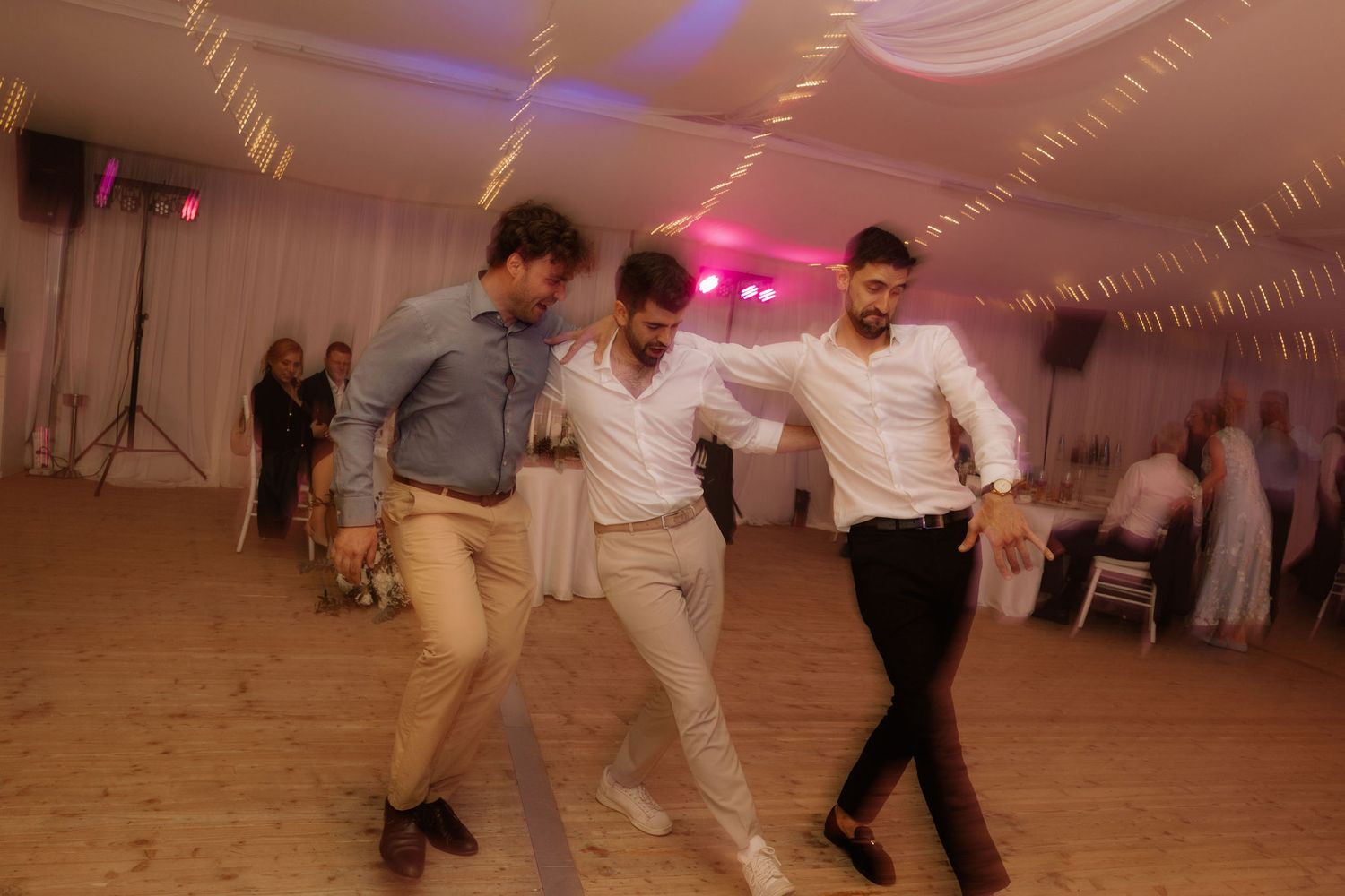 Three men dancing energetically at a wedding reception in a white draped tent venue.