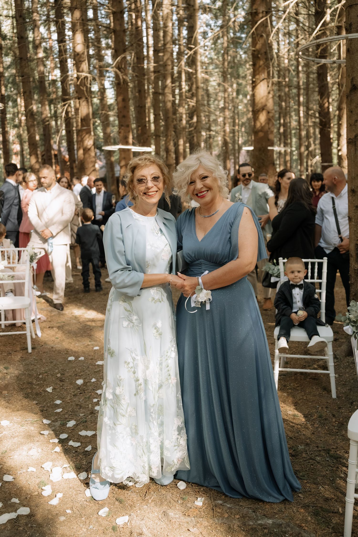 Two elegantly dressed women pose together at an outdoor forest wedding ceremony with guests seated among tall pine trees.